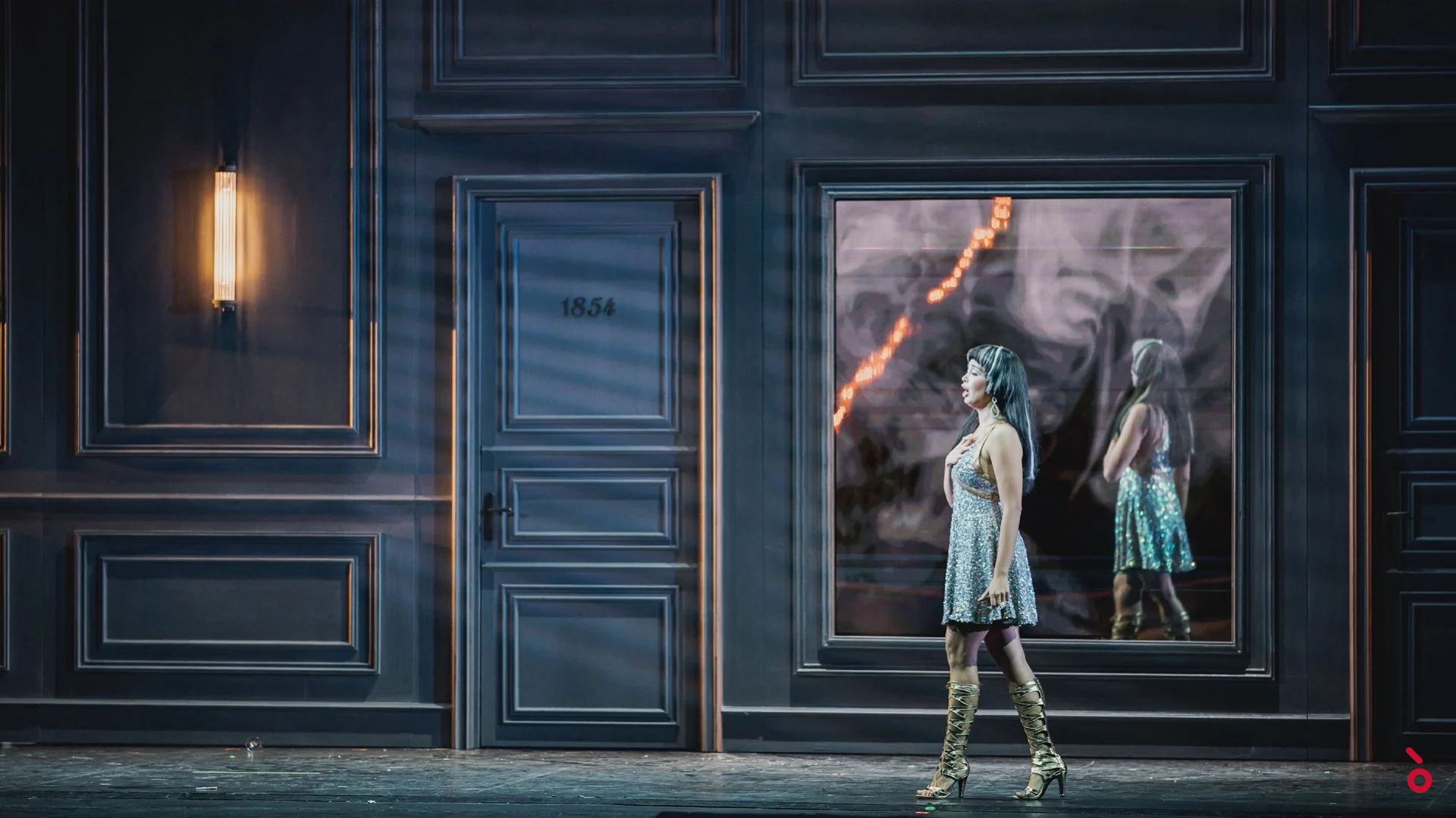 Singer in a sequinned dress and gold boots walking past a numbered door and a mirrored video panel – La Traviata at Maggio Musicale Fiorentino, Florence, Italy, 2022 – opera set design