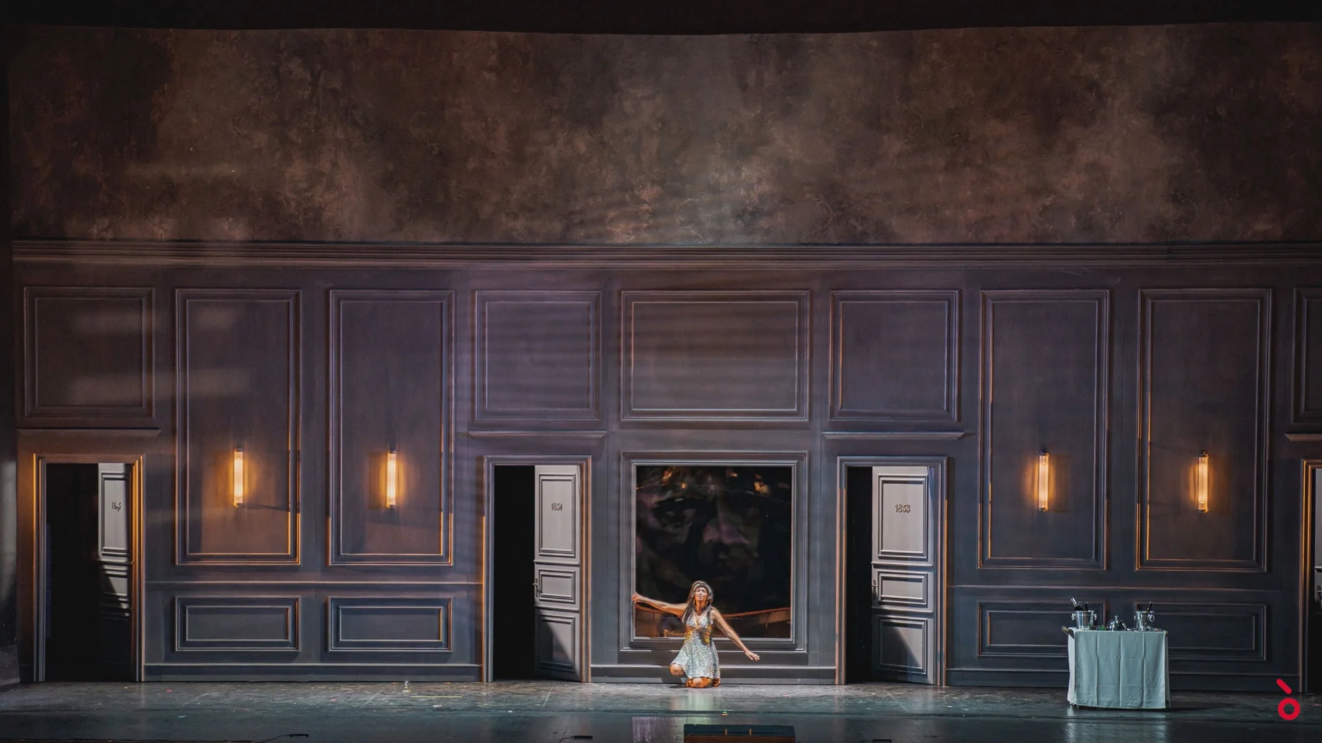 Wide view of the grey panelled set with numbered doors and wall lamps, a woman kneeling at the centre – La Traviata at Maggio Musicale Fiorentino, Florence, Italy, 2022 – opera set design