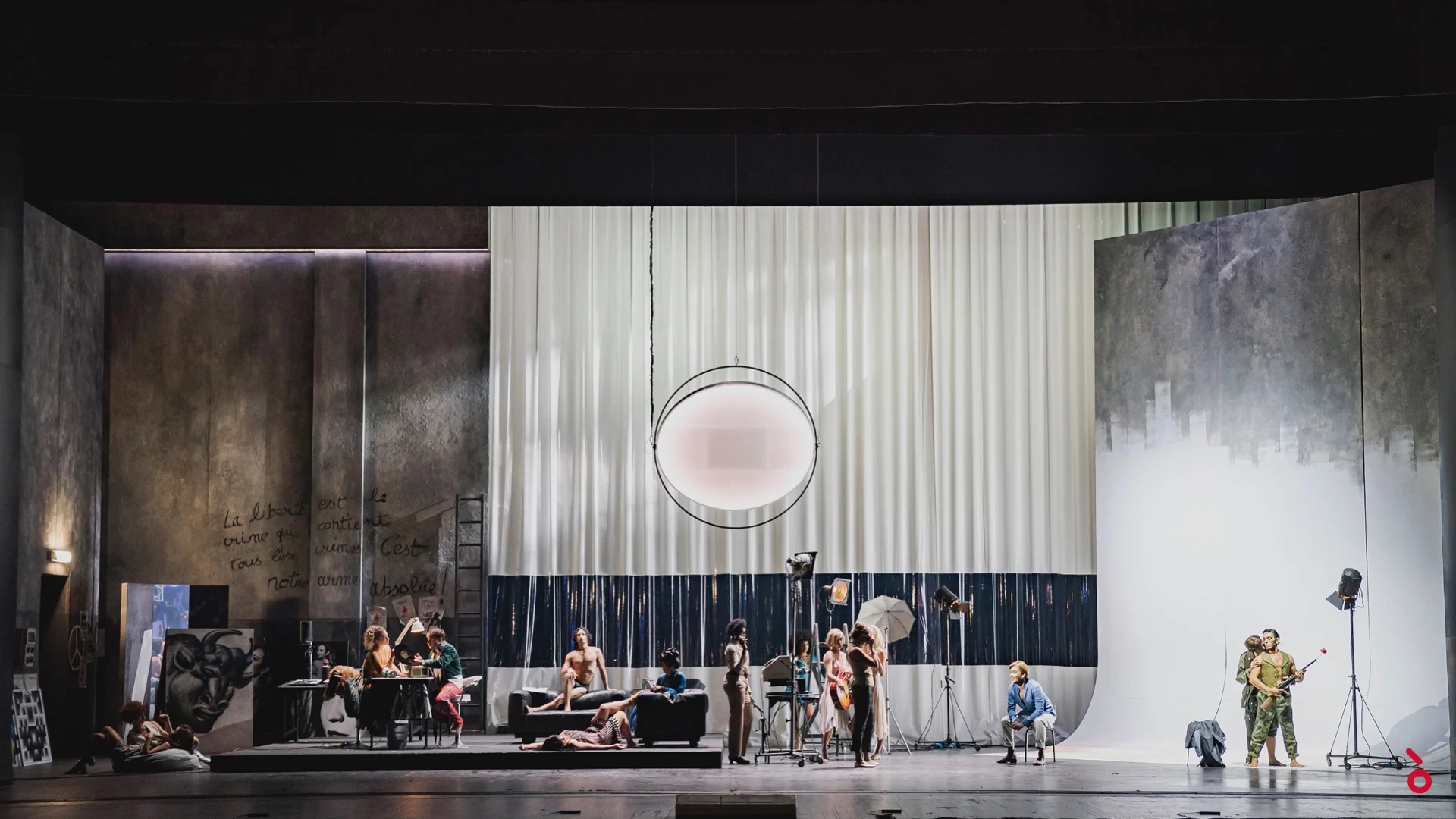 Wide view of a photo studio set with white drapes, a round light and performers posing on sofas – La Traviata at Maggio Musicale Fiorentino, Florence, Italy, 2022 – opera set design