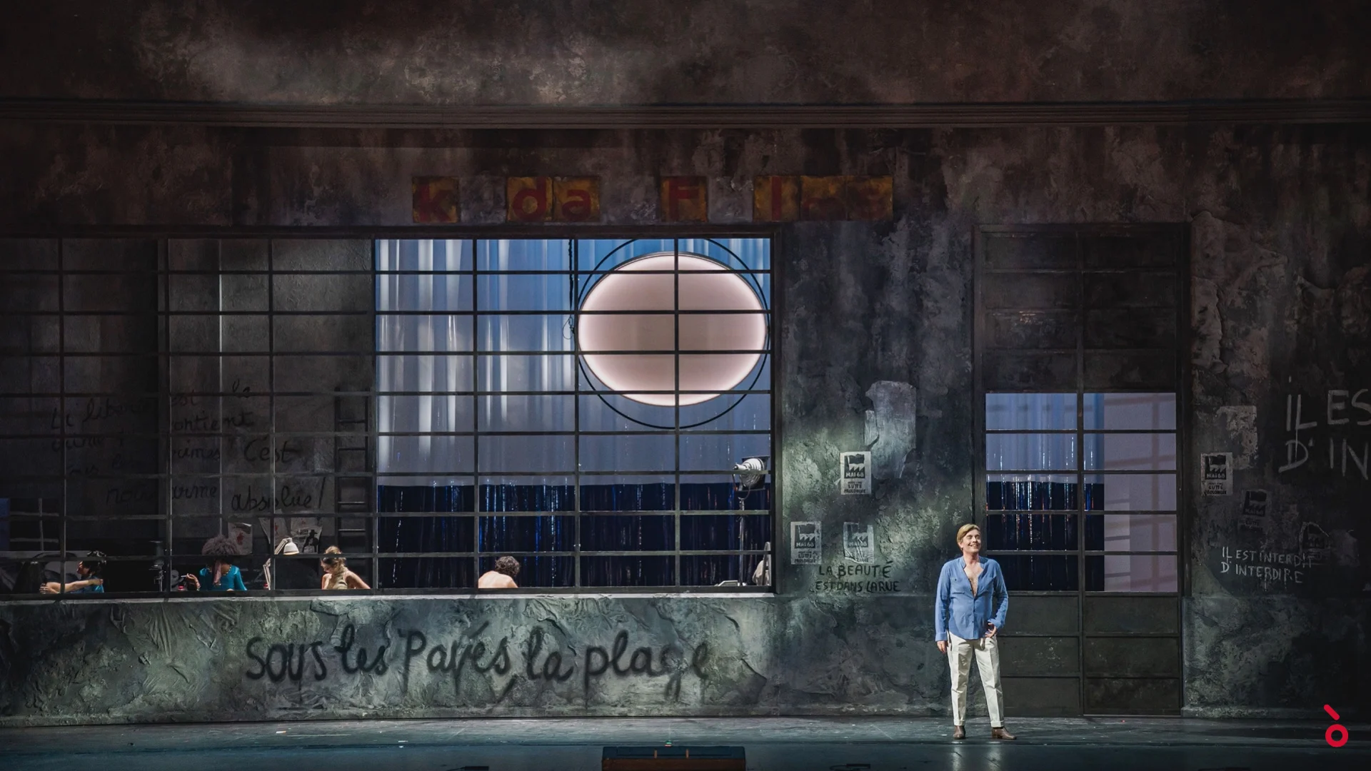 Weathered wall with large windows and the graffiti "Sous les pavés la plage", a singer standing alone in front – La Traviata at Maggio Musicale Fiorentino, Florence, Italy, 2022 – opera set design