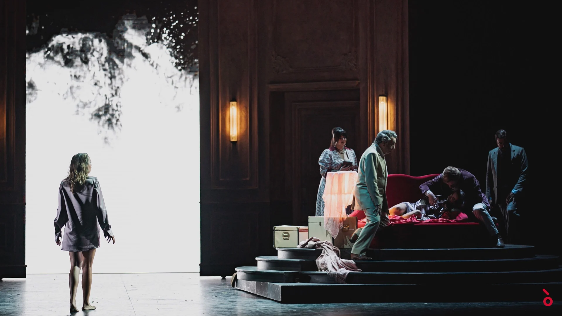 A woman in a shirt facing a glowing white doorway while others gather around a red sofa on stepped platforms – La Traviata at Maggio Musicale Fiorentino, Florence, Italy, 2022 – opera set design