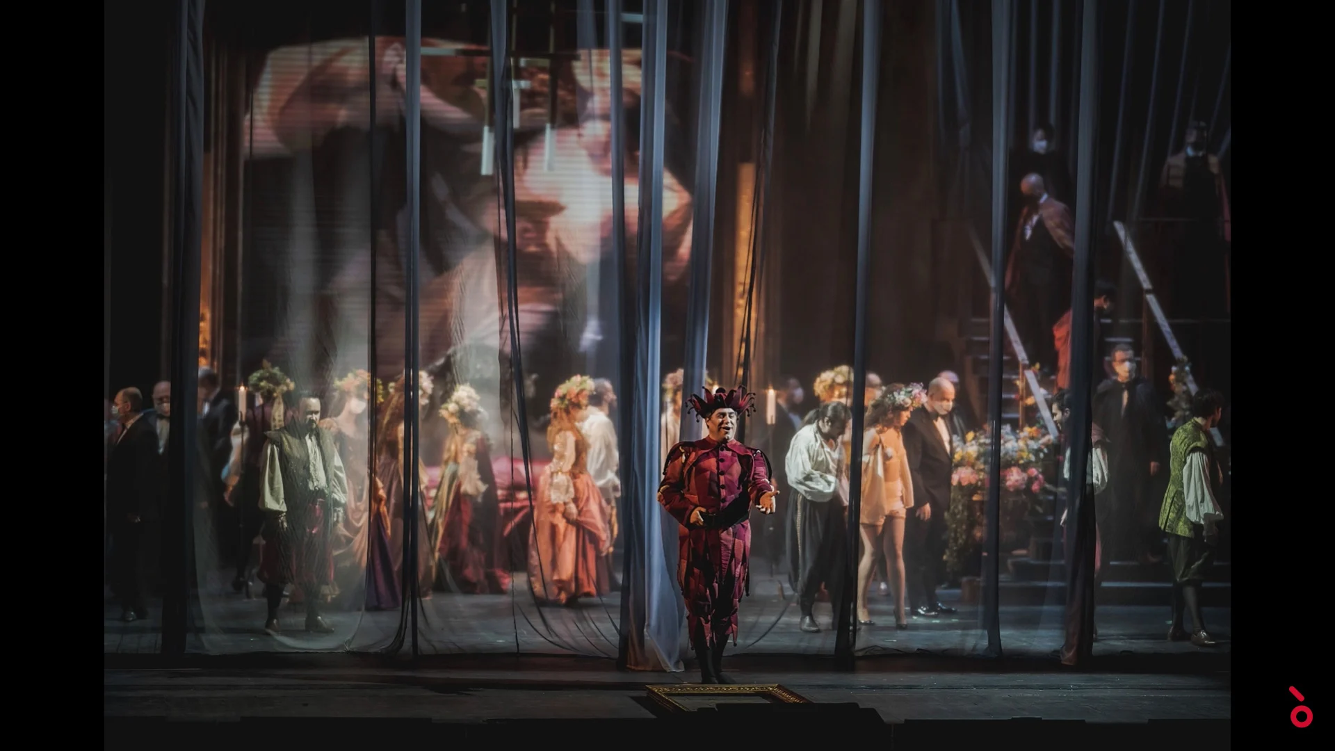 Jester in a red costume before sheer curtains, courtiers behind and a large projected face – Rigoletto at Maggio Musicale Fiorentino, Florence, Italy, 2021 – opera set design