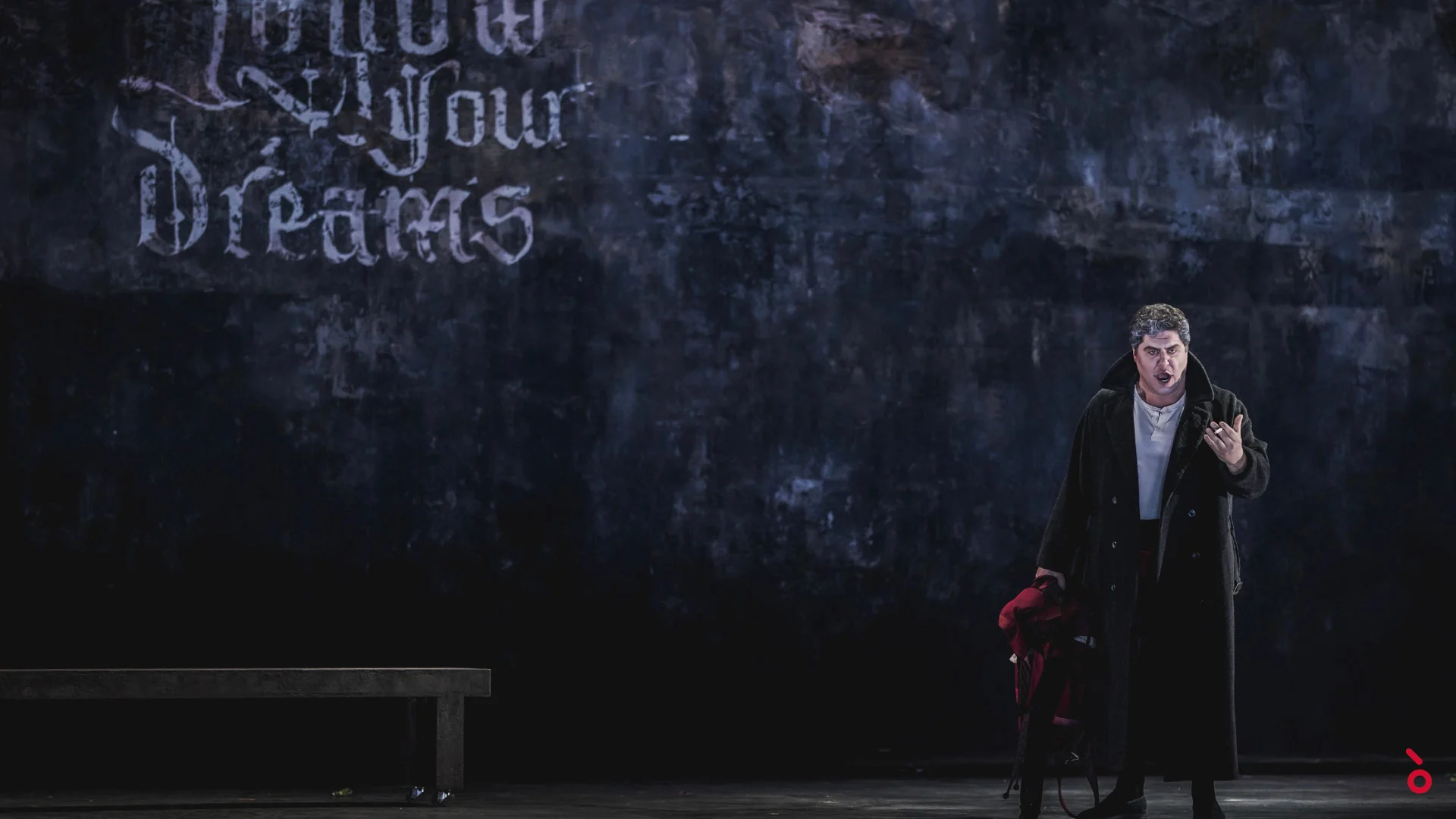 Singer in a long black coat before a dark wall with the graffiti Follow Your Dreams, a bench at left – Rigoletto at Maggio Musicale Fiorentino, Florence, Italy, 2021 – opera set design