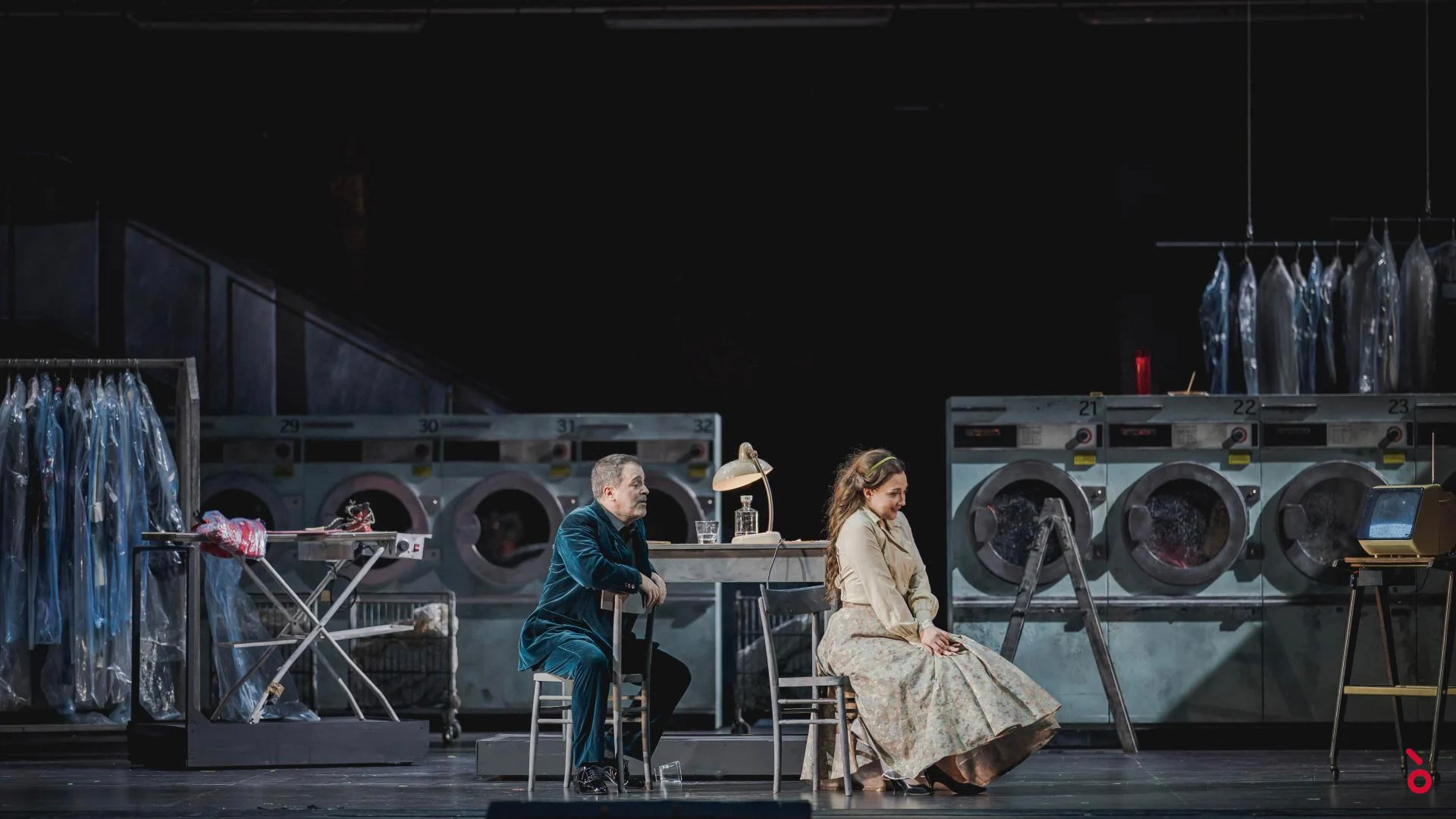 Two singers seated at a table in the laundromat set, washing machines and wrapped clothes behind – Rigoletto at Maggio Musicale Fiorentino, Florence, Italy, 2021 – opera set design