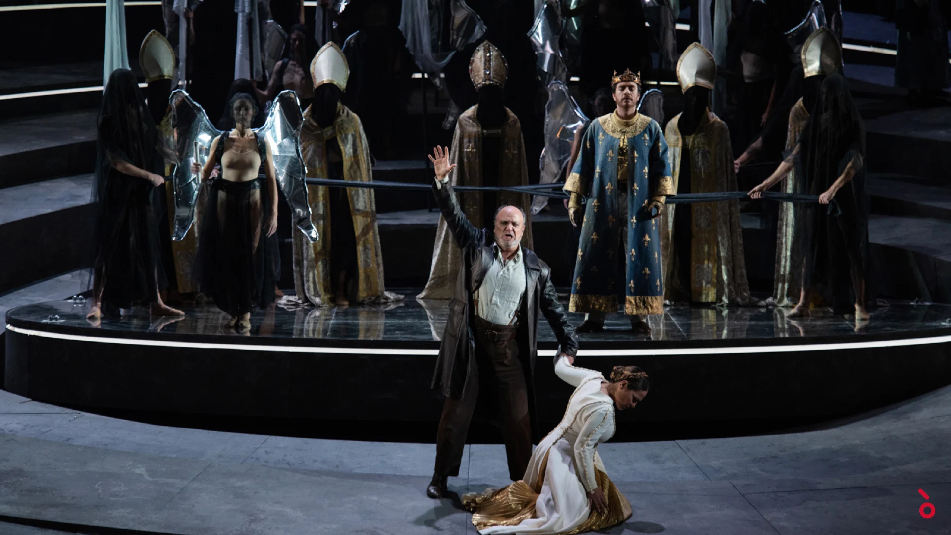 Singer in a leather coat on the circular stage, a woman kneeling at his feet, chorus in mitres behind – Giovanna d'Arco at La Scala, Milan, Italy, 2021 – opera set design