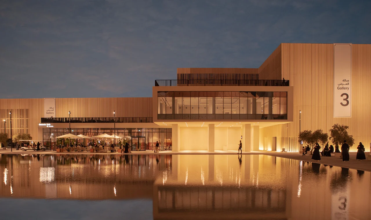 Gallery 3 pavilion at dusk, its warm-lit slatted facade and glazed upper floor reflected in a long pool – Islamic Arts Biennale, Jeddah, Saudi Arabia, 2023 – architecture
