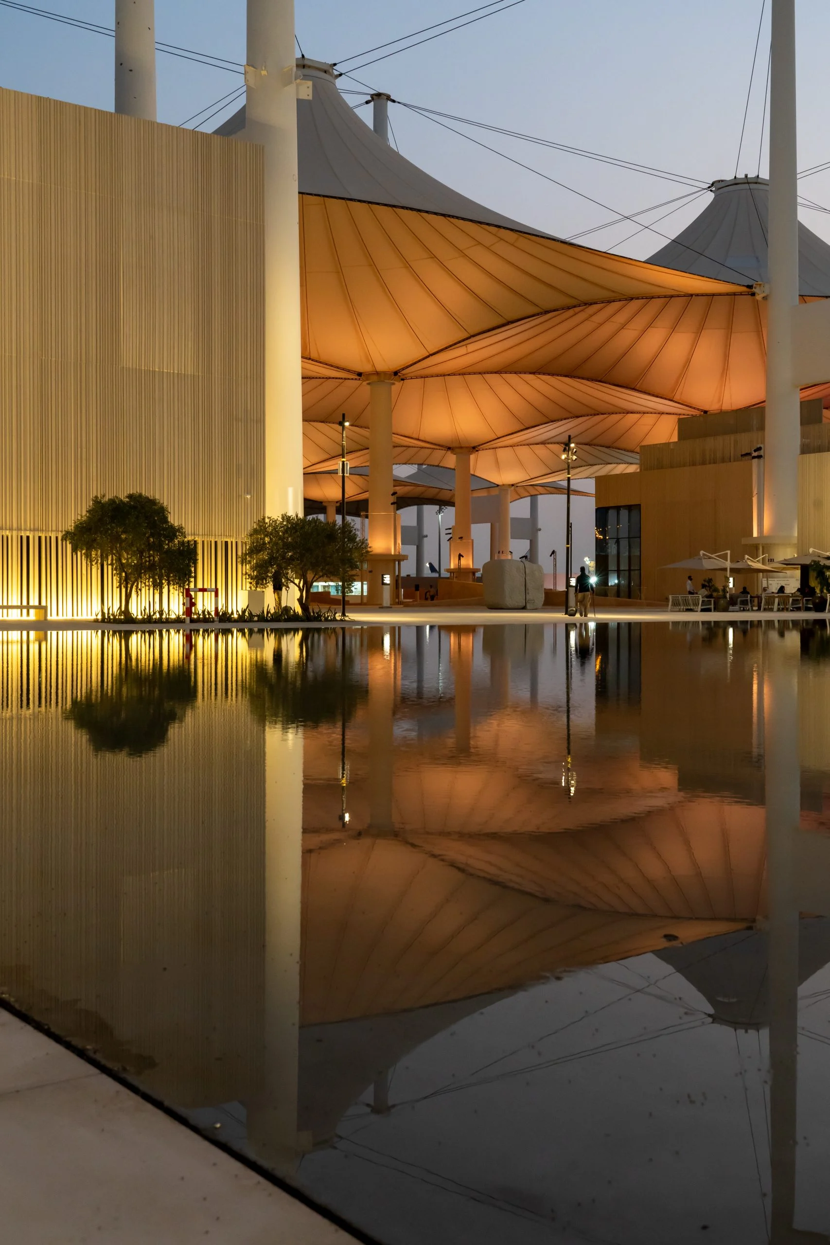 Hajj Terminal fabric canopies glowing at dusk beside a slatted pavilion, reflected in still water – Islamic Arts Biennale, Jeddah, Saudi Arabia, 2023 – architecture