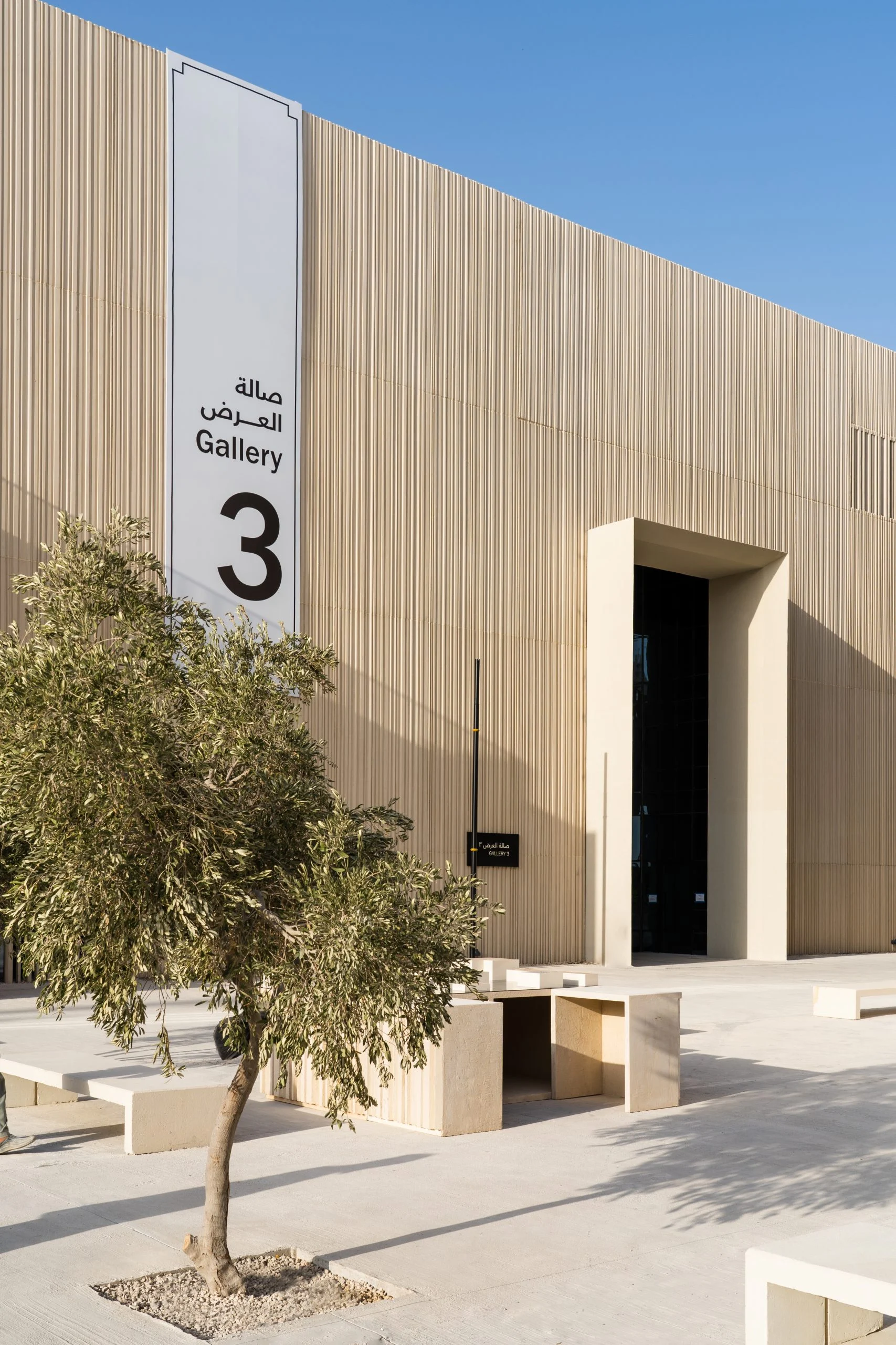Slatted beige facade of Gallery 3 with bilingual signage, an olive tree and stone benches in the sun – Islamic Arts Biennale, Jeddah, Saudi Arabia, 2023 – architecture