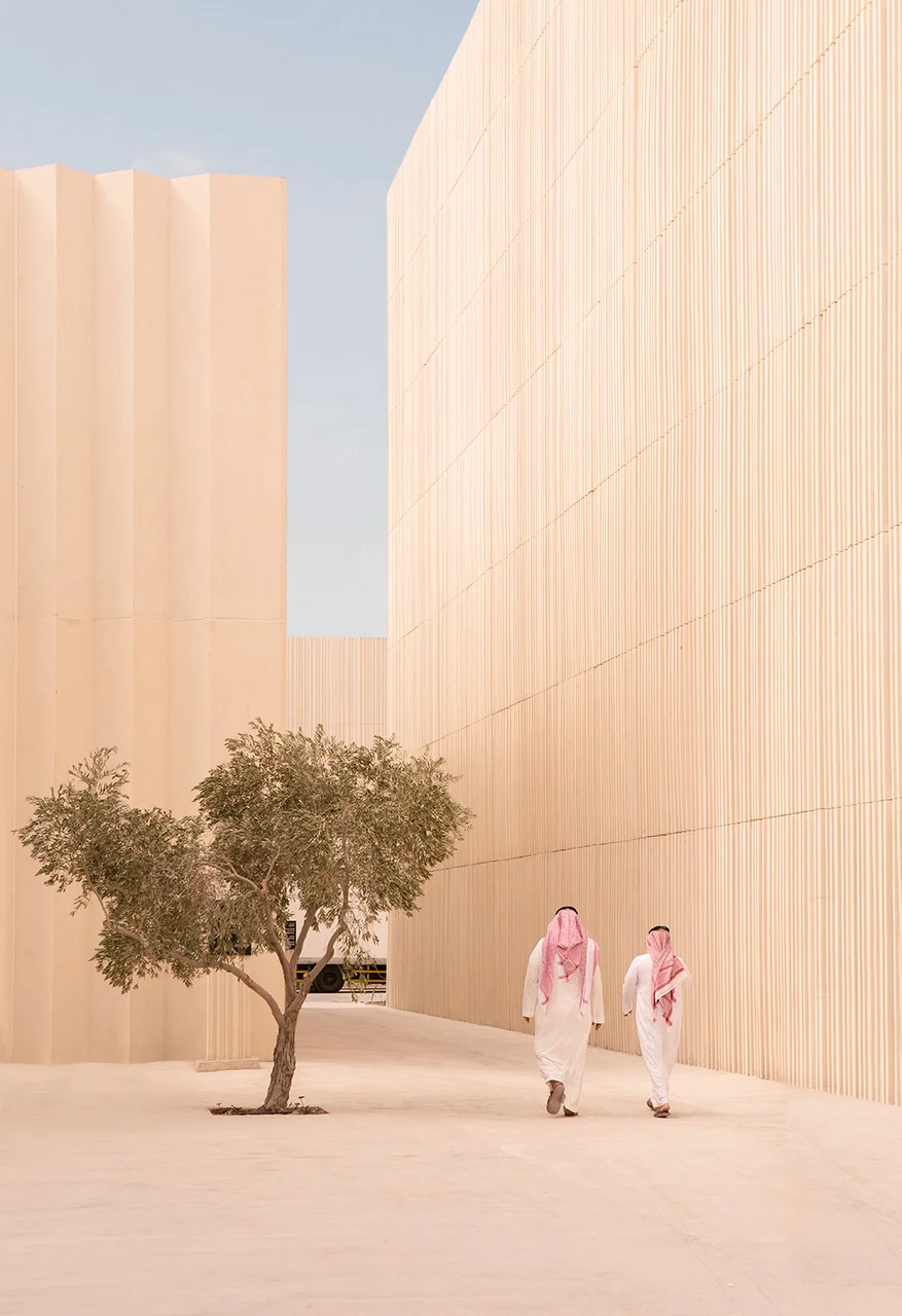 Two visitors in white thobes walking past an olive tree between tall pale slatted pavilion walls – Islamic Arts Biennale, Jeddah, Saudi Arabia, 2023 – architecture