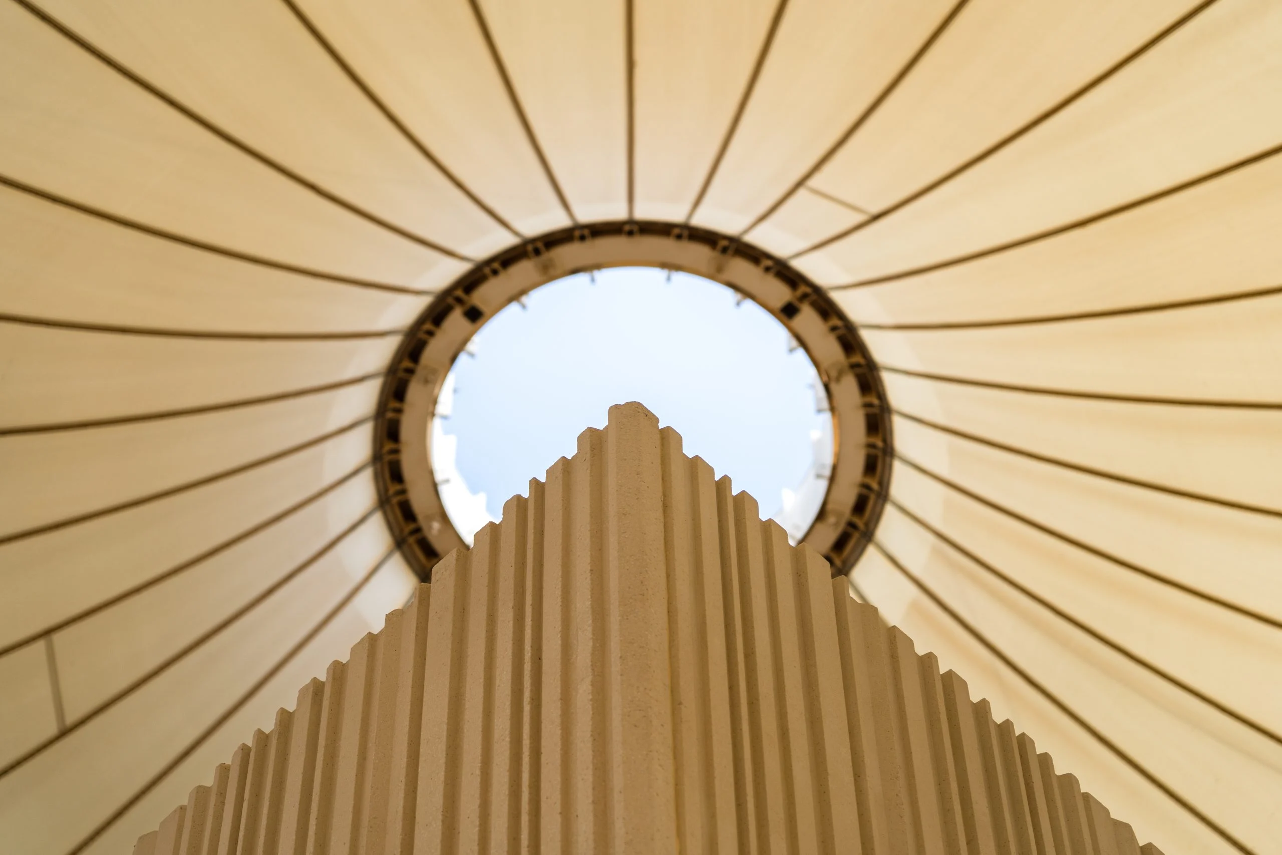 Upward view of a ribbed sculptural column rising toward the circular oculus of a fabric tent roof – Islamic Arts Biennale, Jeddah, Saudi Arabia, 2023 – architecture
