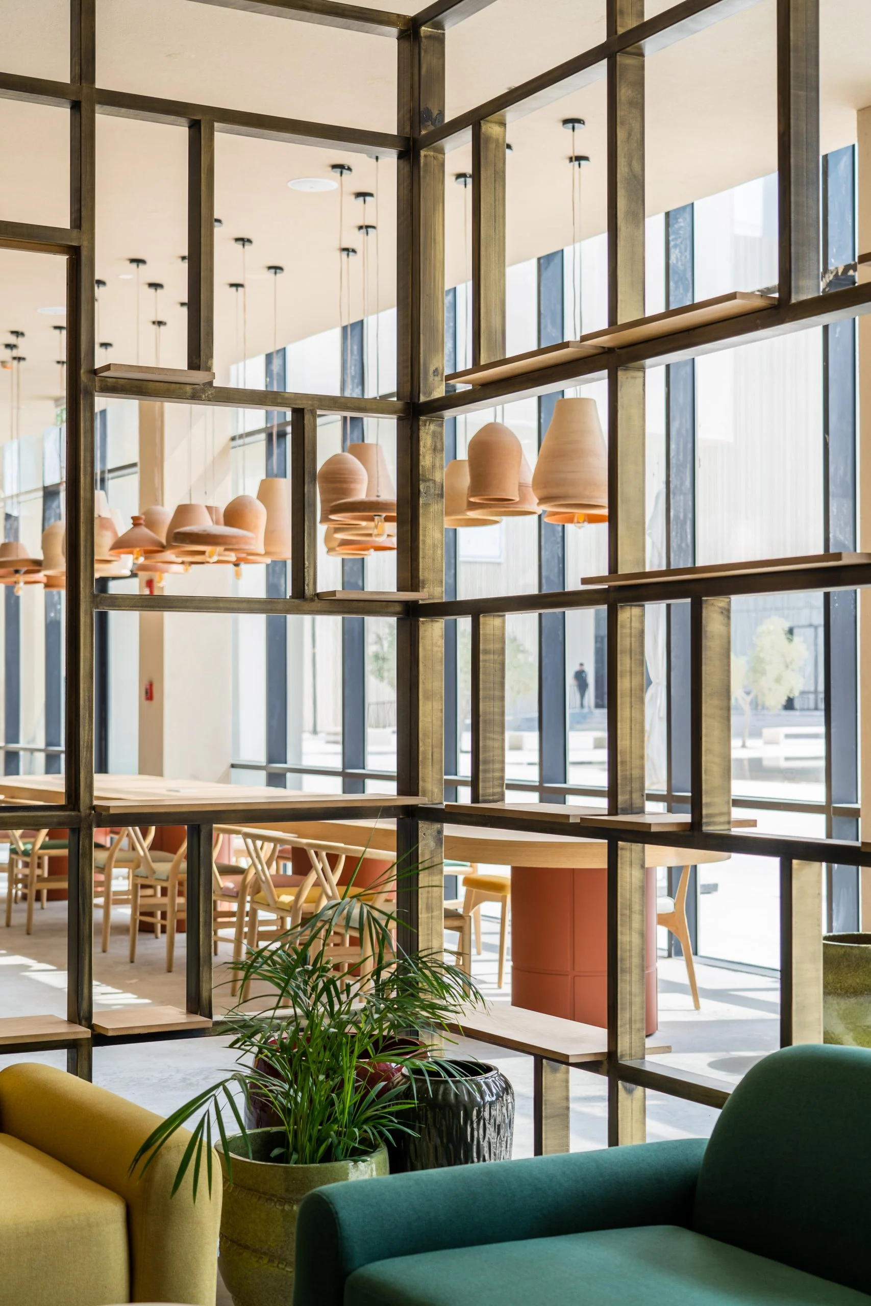 Brass grid shelving dividing the cafe, with terracotta pendants, potted palms and green armchairs – Islamic Arts Biennale, Jeddah, Saudi Arabia, 2023 – architecture