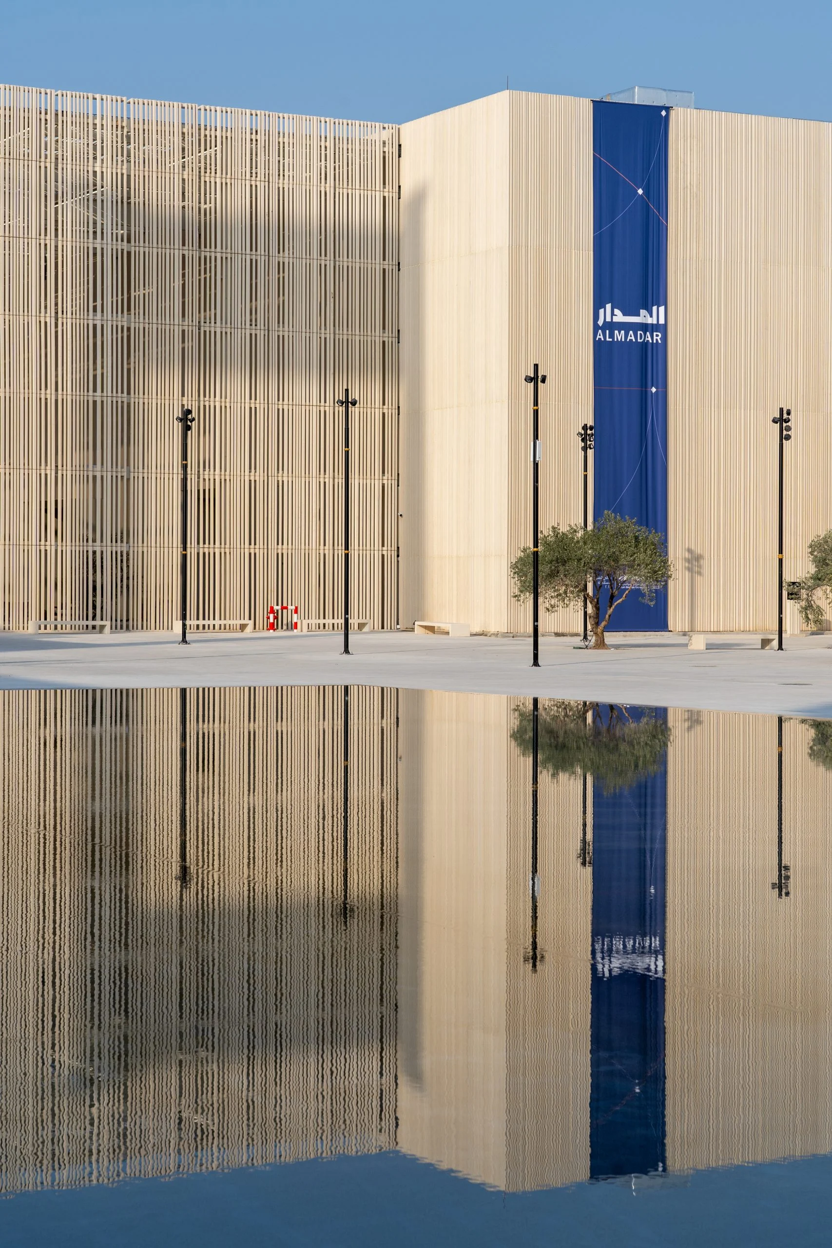 Slatted pavilion facade with a blue AlMadar banner, mirrored in a reflecting pool under a clear sky – Islamic Arts Biennale, Jeddah, Saudi Arabia, 2023 – architecture