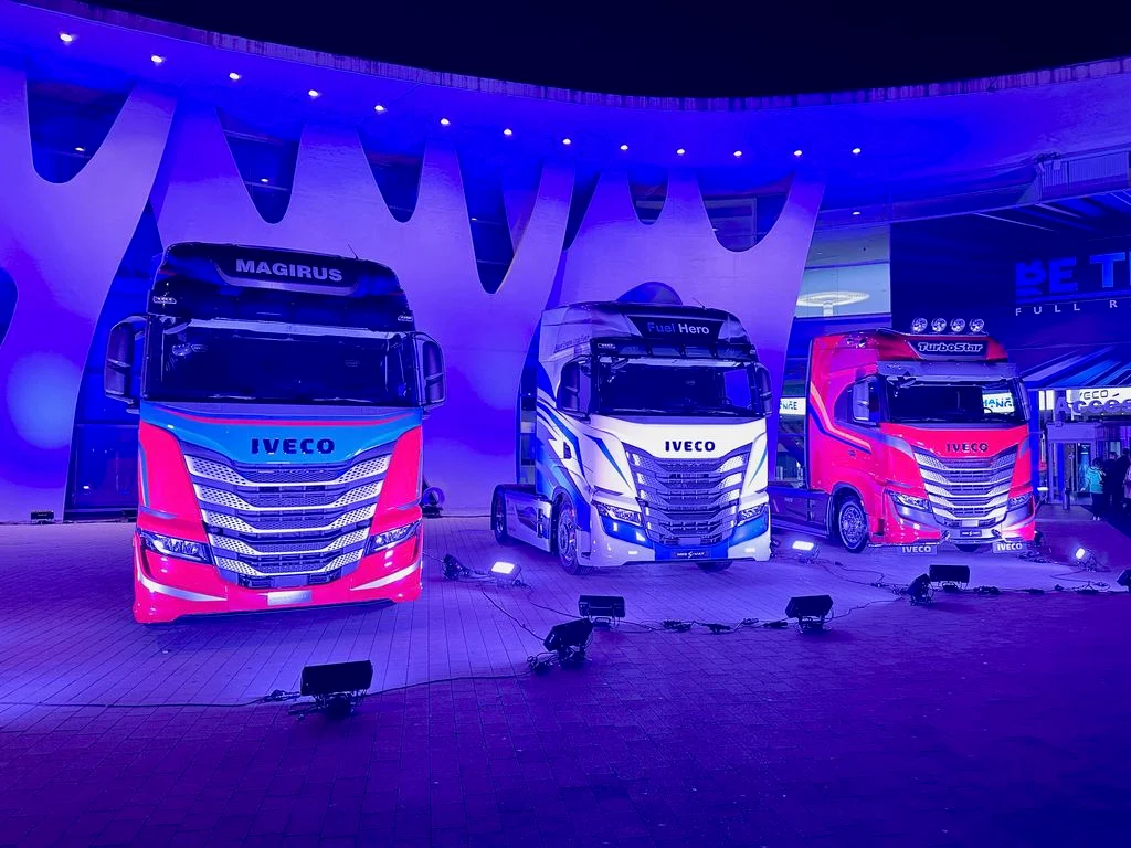 Three truck cabs in pink, white and blue liveries under a wavy canopy lit in purple – IVECO Be The Change Launch, Barcelona, Spain, 2023 – event design