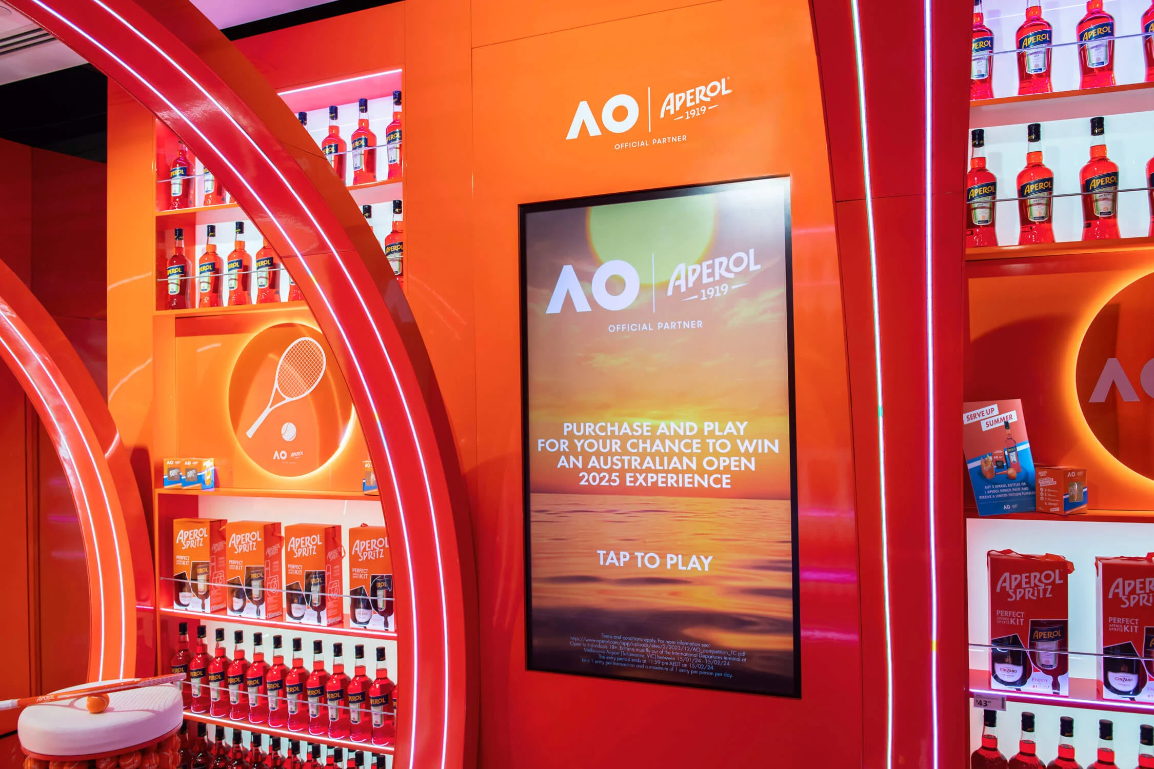 Touchscreen inviting travellers to play for an Australian Open experience, between orange bottle shelves – Aperol Pop-Up at Melbourne Airport, Australia, 2024 – retail design