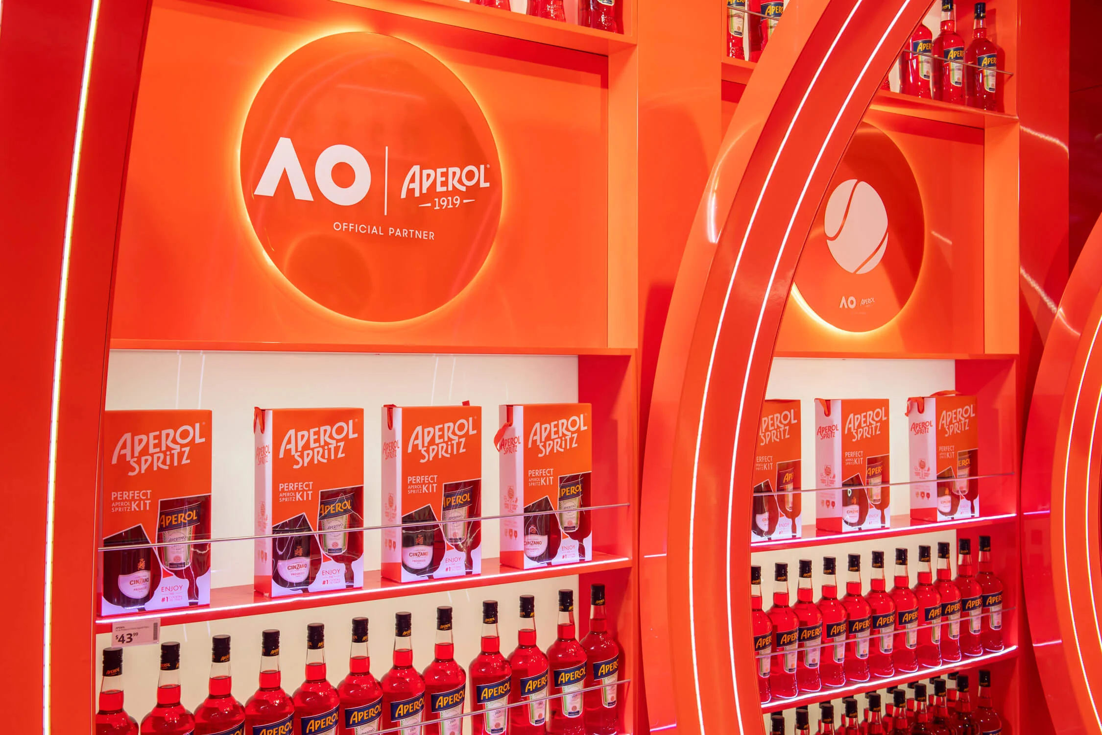 Detail of shelves with Aperol bottles and gift boxes under a backlit Australian Open and Aperol logo – Aperol Pop-Up at Melbourne Airport, Australia, 2024 – retail design