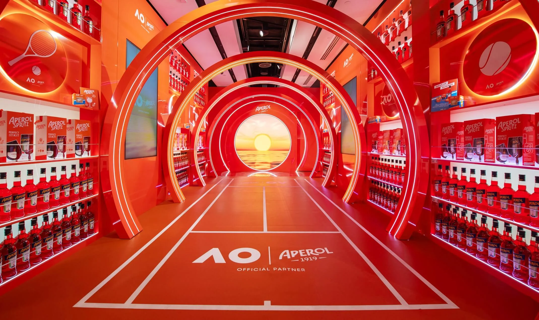 Orange Aperol shop-in-shop: a tunnel of lit arches over a tennis-court floor leading to a sunset screen – Aperol Pop-Up at Melbourne Airport, Australia, 2024 – retail design