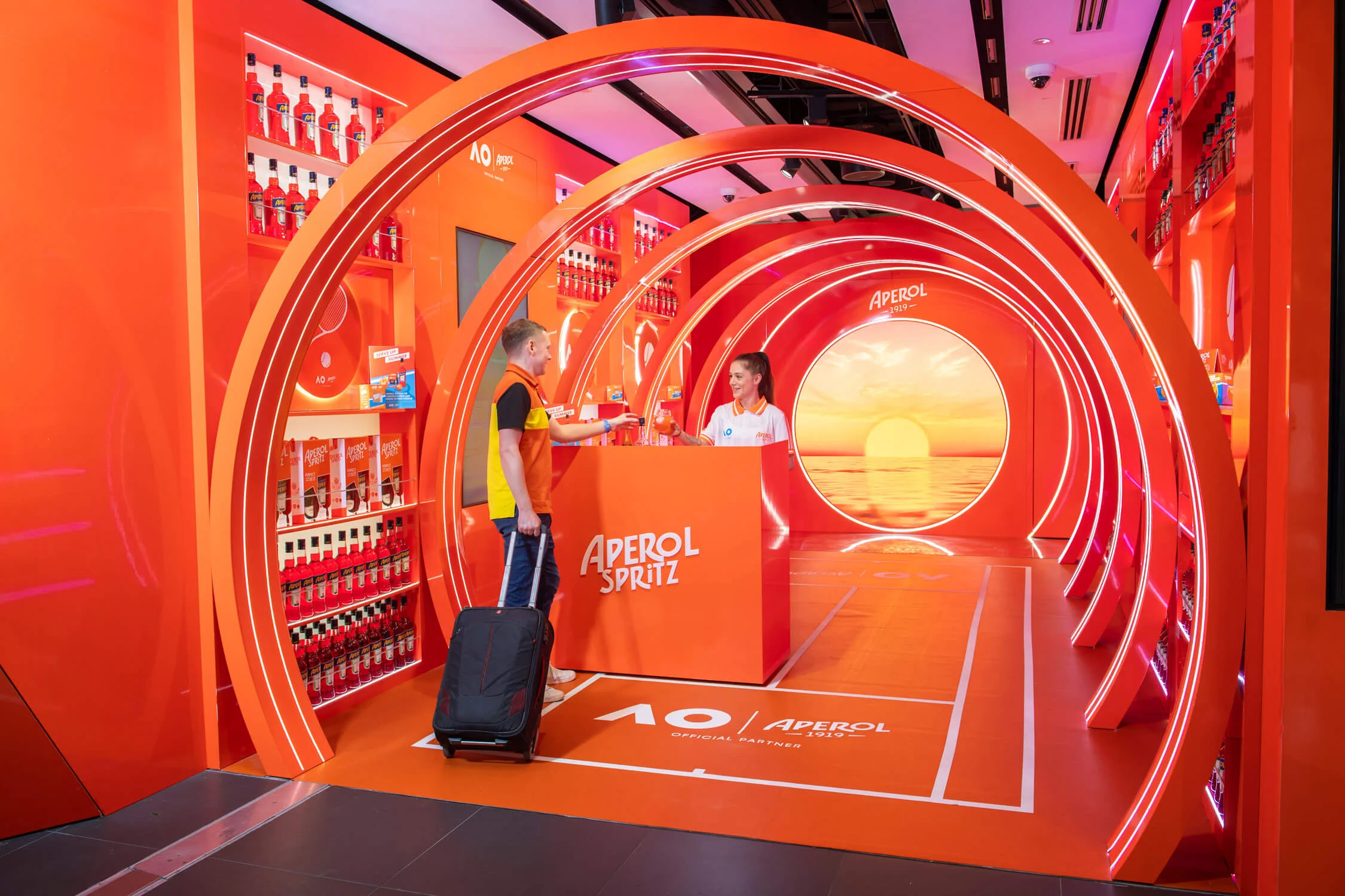 Traveller with a suitcase at the Aperol Spritz counter inside the orange arched tunnel – Aperol Pop-Up at Melbourne Airport, Australia, 2024 – retail design