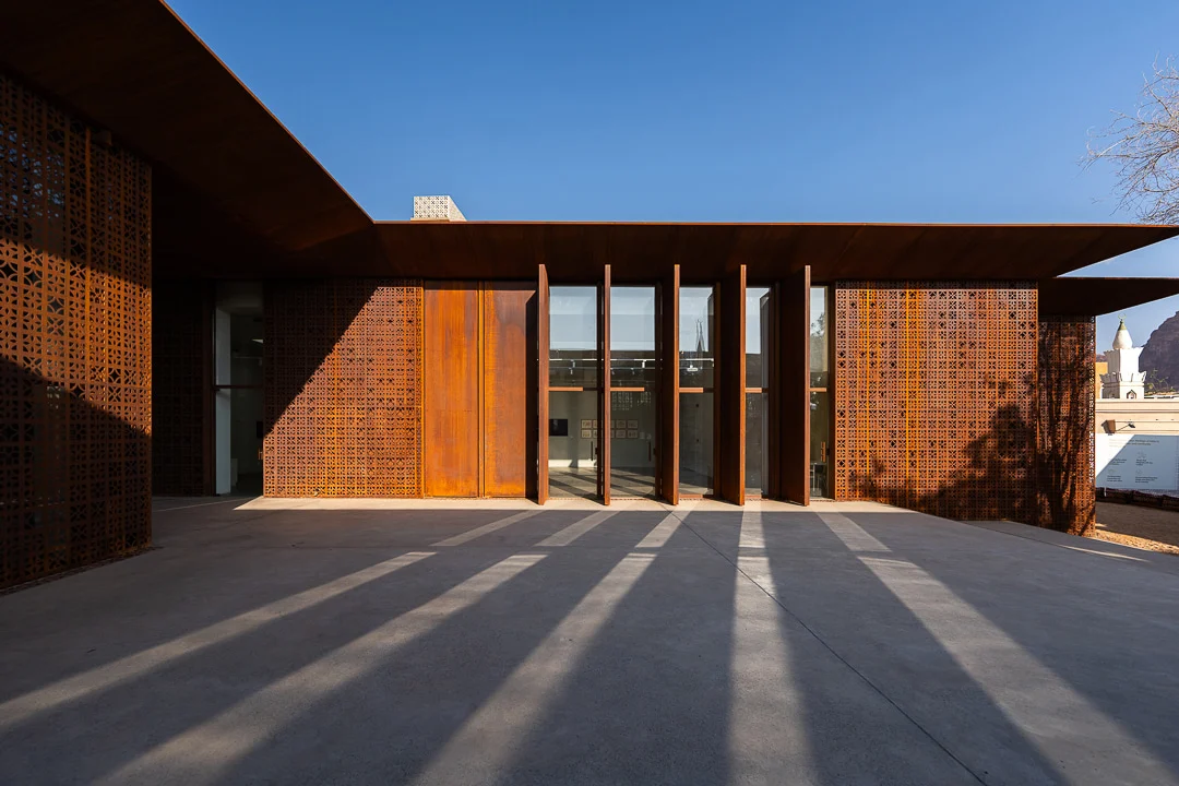 Glazed entrance between rust-coloured fins and perforated screens, long shadows across the concrete forecourt – Design Space AlUla, Saudi Arabia, 2024 – architecture