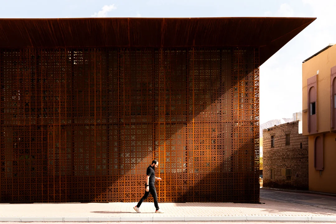 Person walking past the perforated screen facade in raking sunlight, under the deep roof overhang – Design Space AlUla, Saudi Arabia, 2024 – architecture