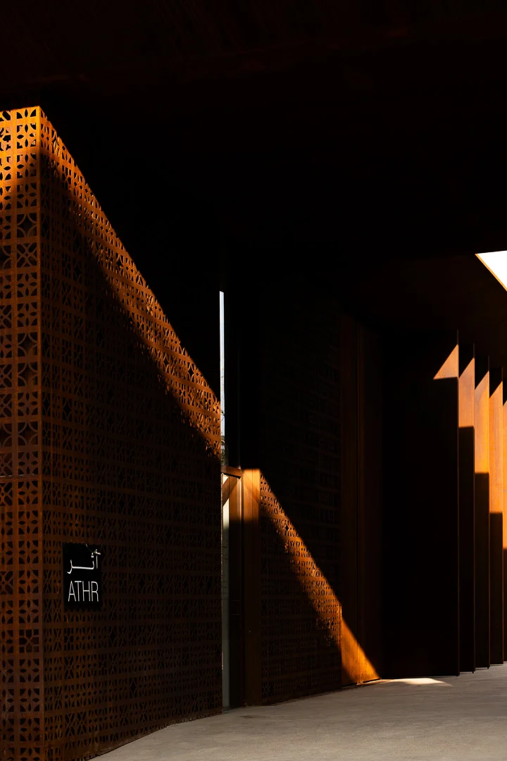 Shadowed corner of the perforated screen with an ATHR sign in Arabic and English, light slanting across – Design Space AlUla, Saudi Arabia, 2024 – architecture