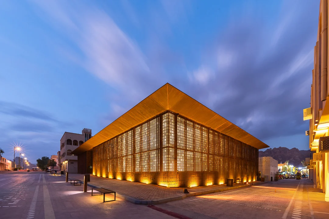 Design Space AlUla at dusk: glowing perforated metal facade under a deep overhanging roof, seen from the street – Design Space AlUla, Saudi Arabia, 2024 – architecture