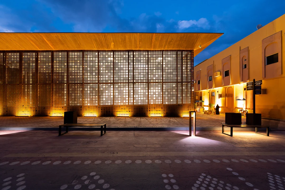 Front elevation at dusk, the perforated screen lit from within beside a yellow neighbouring building – Design Space AlUla, Saudi Arabia, 2024 – architecture