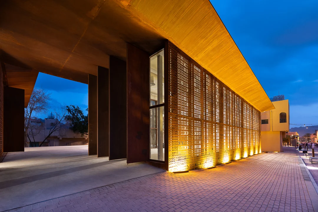 Side view at dusk of the lit perforated screen and deep roof overhang above a covered passage – Design Space AlUla, Saudi Arabia, 2024 – architecture