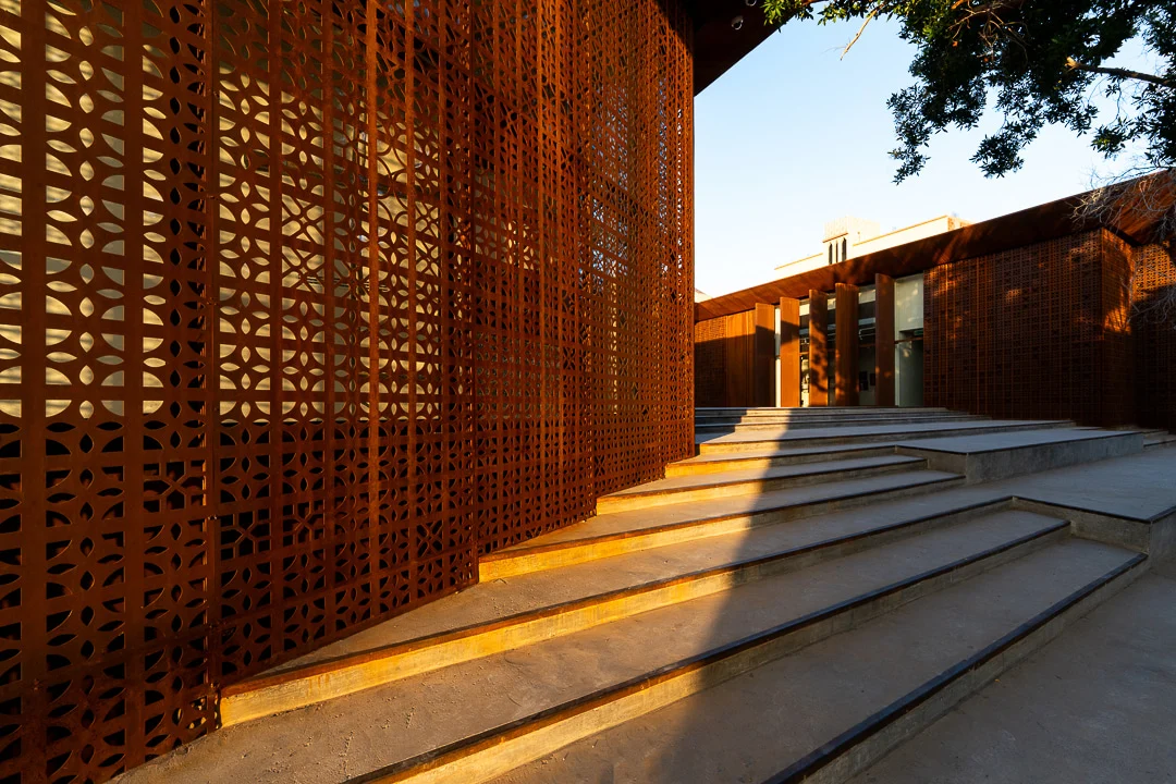 Wide concrete steps alongside the sunlit perforated screen, leading to the covered entrance – Design Space AlUla, Saudi Arabia, 2024 – architecture
