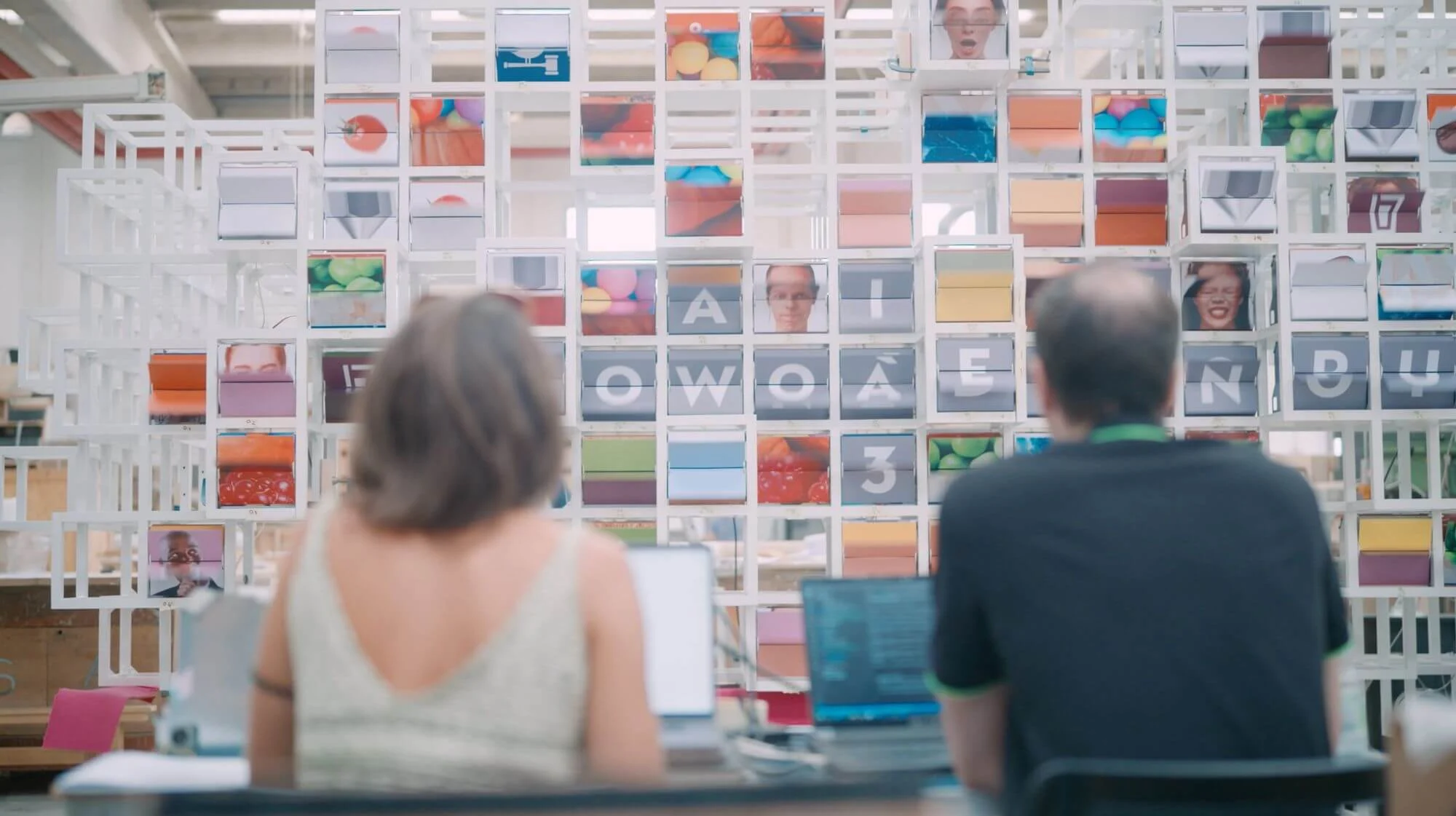 Two people at laptops facing La Nuvola during testing, its modules showing letters, faces and colours – La Nuvola at Cirfood District, Reggio Emilia, Italy, 2022 – kinetic installation design