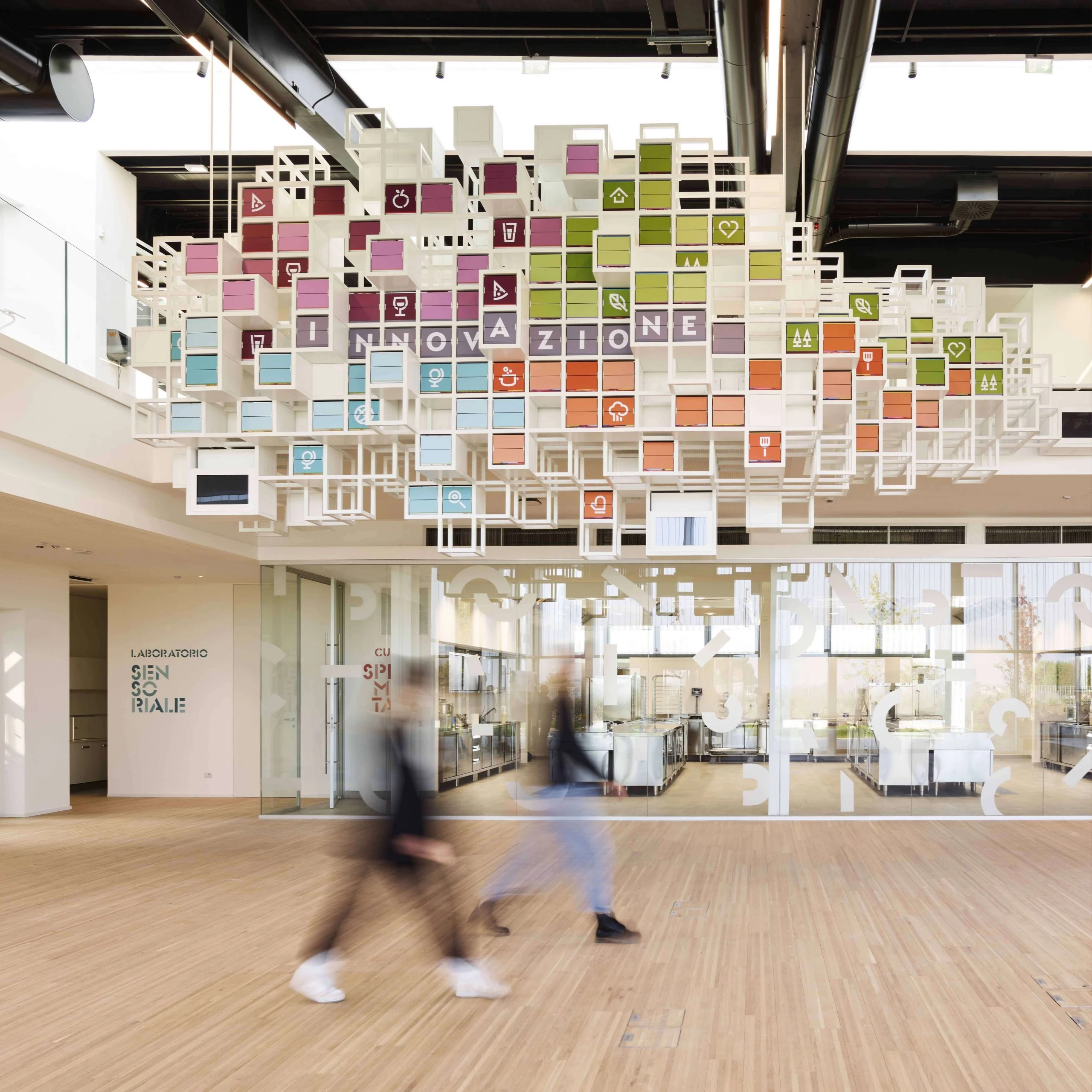 Visitors walking beneath La Nuvola, a suspended cloud of colourful split-flap cubes spelling "Innovazione" – La Nuvola at Cirfood District, Reggio Emilia, Italy, 2022 – kinetic installation design
