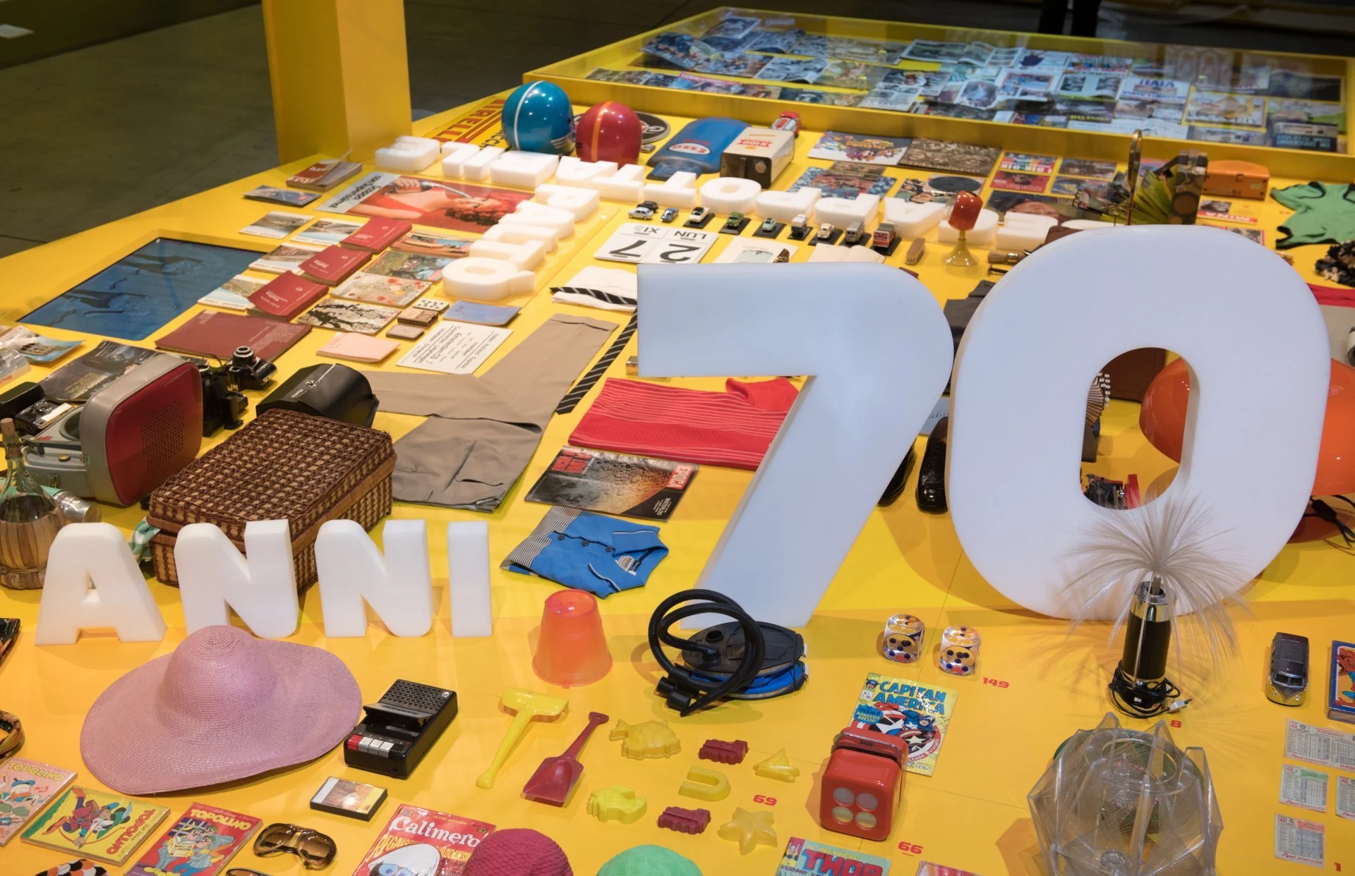 Detail of the yellow display table with the words Anni 70 among hats, toys, clothes and objects of the 1970s – Esselunga Supermostra 60th Anniversary, Milan, Italy, 2018 – exhibition design