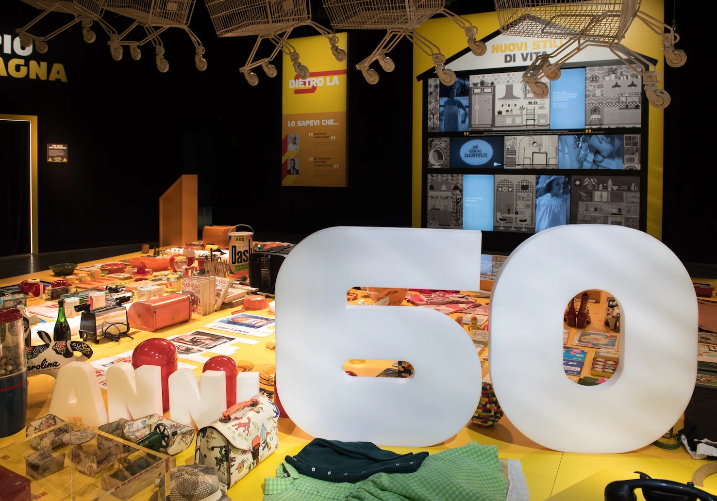 Large white 60 letters on a yellow table covered with everyday objects, with hanging trolleys above – Esselunga Supermostra 60th Anniversary, Milan, Italy, 2018 – exhibition design