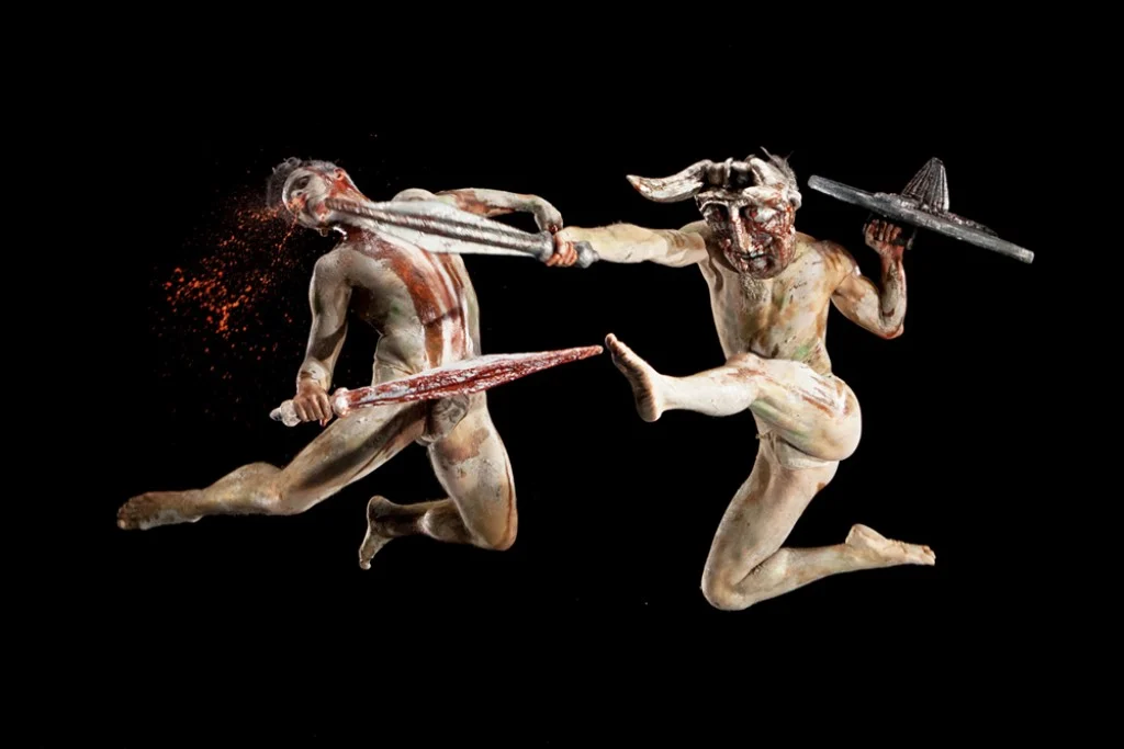 Two clay-painted warriors with sword and shield clash in mid-air, one wearing a horned helmet – I Shardana at Teatro Lirico di Cagliari, Italy, 2013 – opera set design