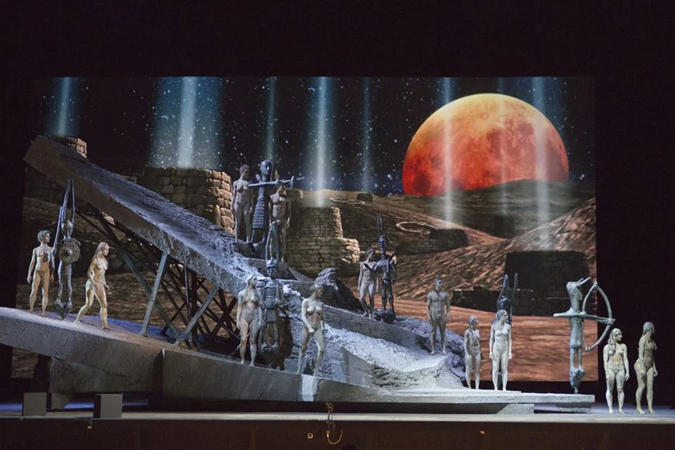 Clay-covered performers on a tilted rock set before a projected desert landscape and huge red moon – I Shardana at Teatro Lirico di Cagliari, Italy, 2013 – opera set design