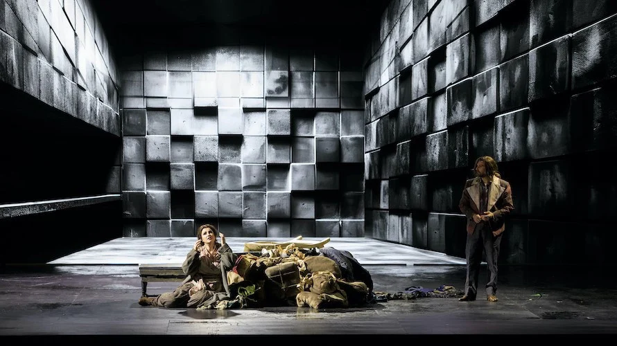 Soprano seated beside a pile of sacks in a stark room of grey stone blocks, a singer watching from the right – Il Trovatore at Teatro Comunale Nouveau, Bologna, Italy, 2024 – opera set design