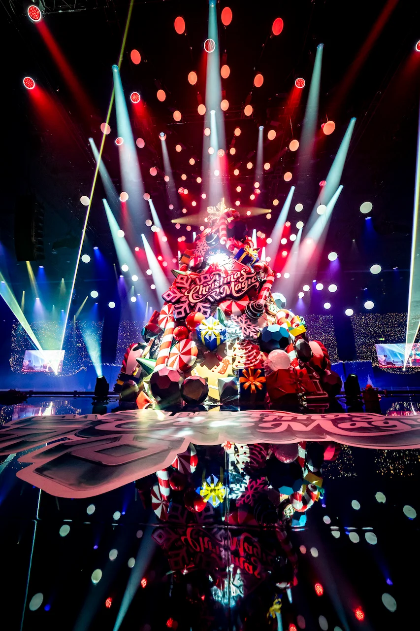Giant Christmas tree of sweets and gifts under radiating spotlights, reflected in a glossy floor – A Christmas Magic at Allianz MiCo, Milan, Italy, 2023 – event design