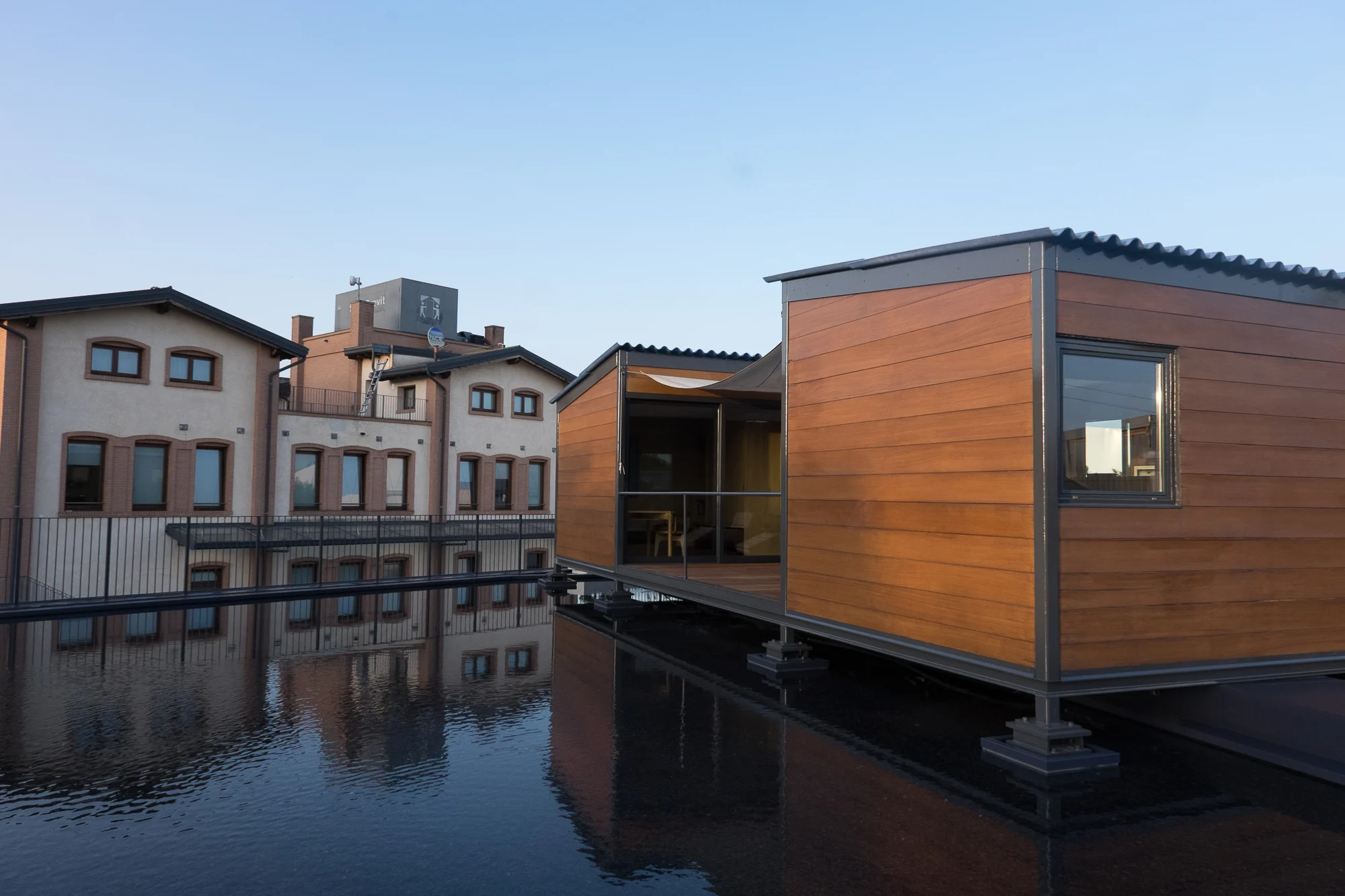 Wood-clad pavilion with corrugated roof on stilts over a reflecting pool, next to the brick factory buildings – Factory Reflections at Sice Previt, Segrate, Italy, 2024 – exhibition design
