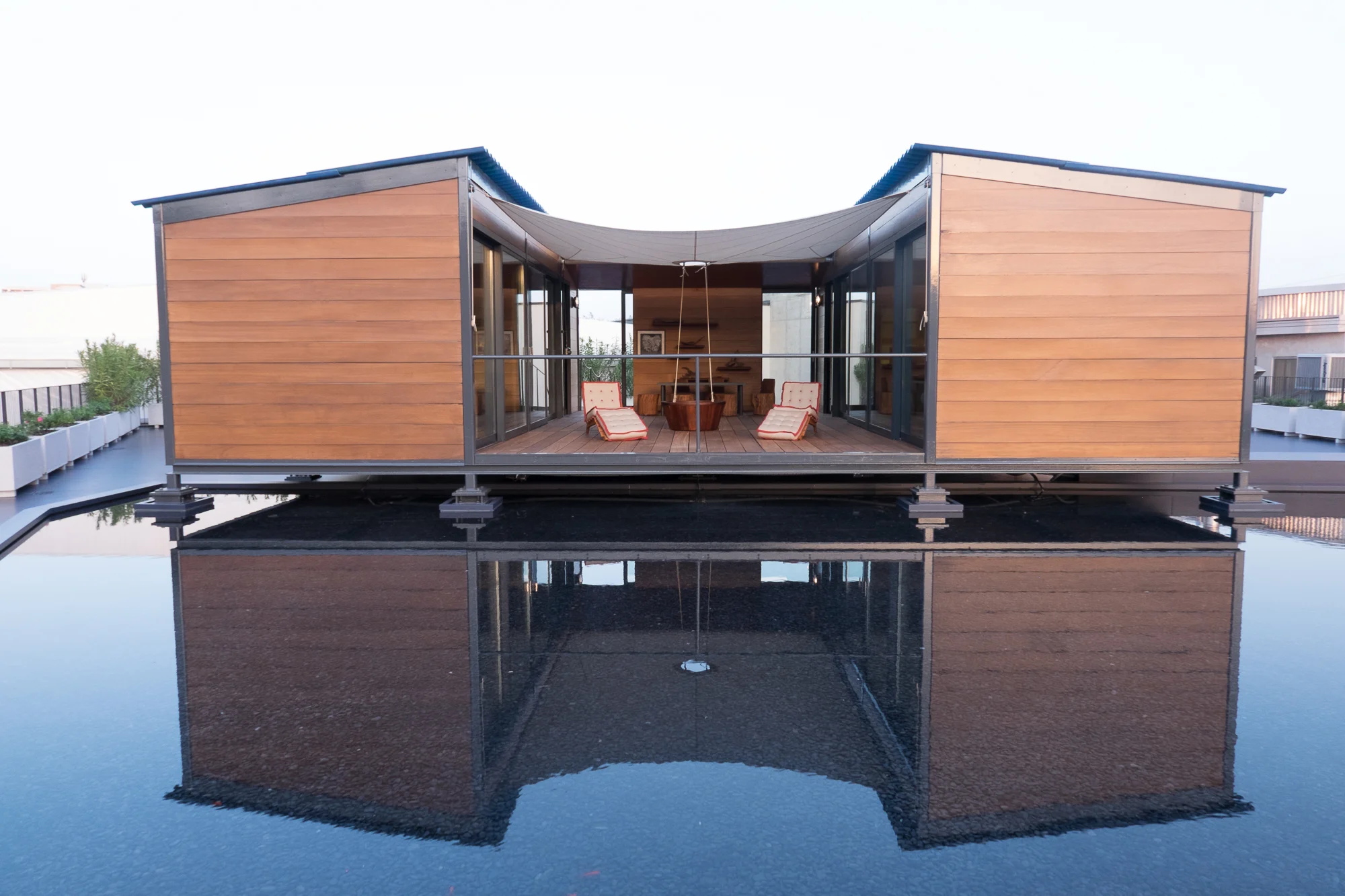 Symmetrical view of the wood-clad pavilion with an open central lounge, mirrored in the rooftop water basin – Factory Reflections at Sice Previt, Segrate, Italy, 2024 – exhibition design