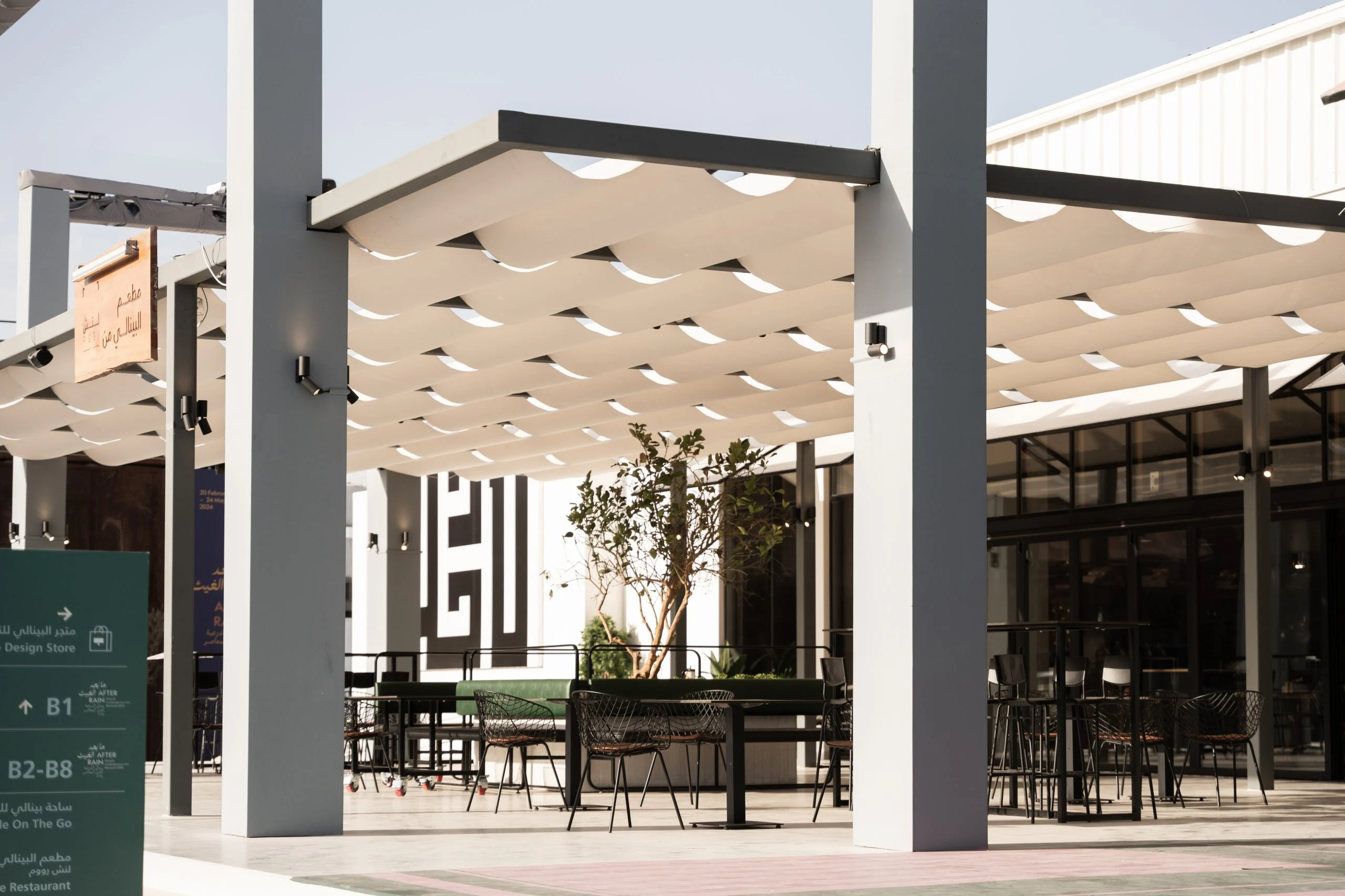 Outdoor seating under white pleated fabric canopies on slender columns, a kufic-style graphic behind – Diriyah Contemporary Art Biennale, Riyadh, Saudi Arabia, 2024 – architecture
