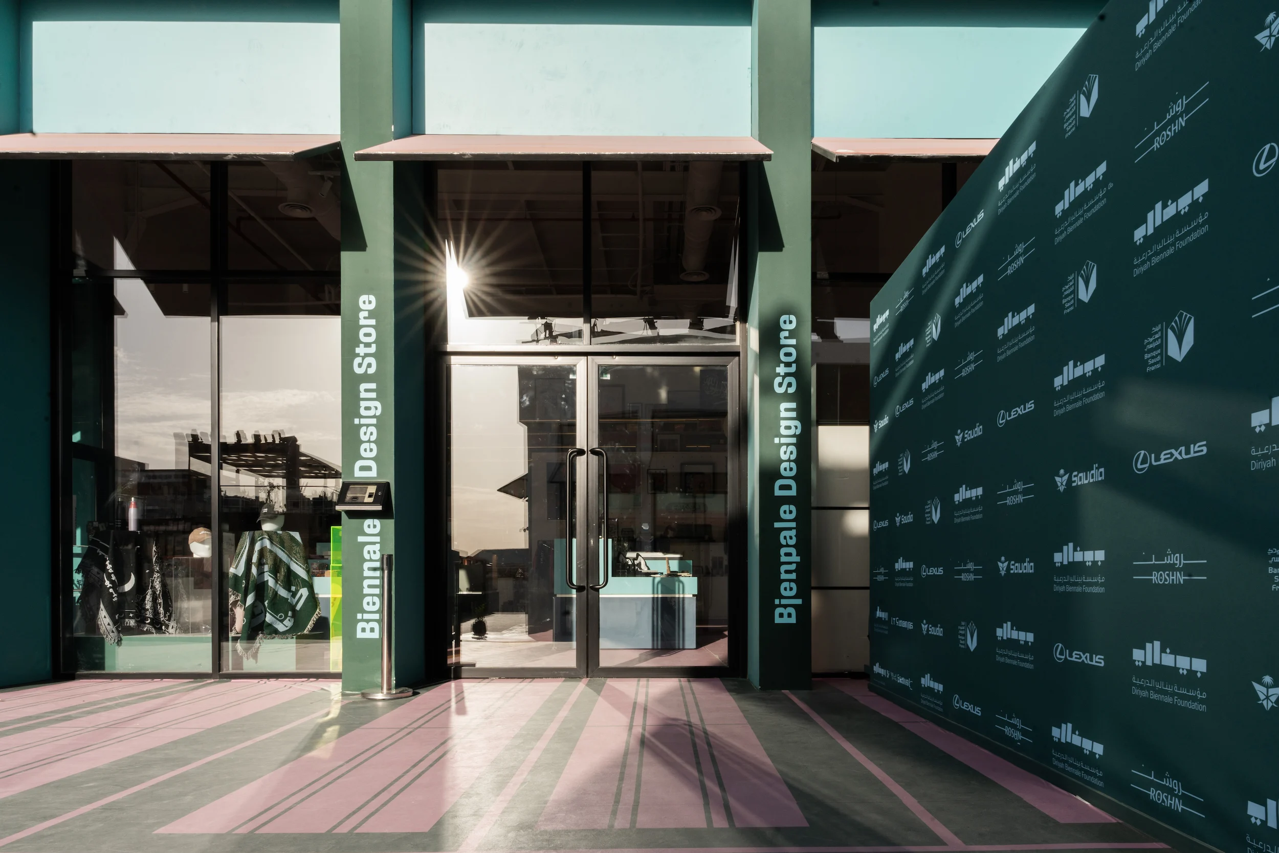 Green entrance of the Biennale Design Store beside a sponsor wall, pink and green striped paving – Diriyah Contemporary Art Biennale, Riyadh, Saudi Arabia, 2024 – architecture