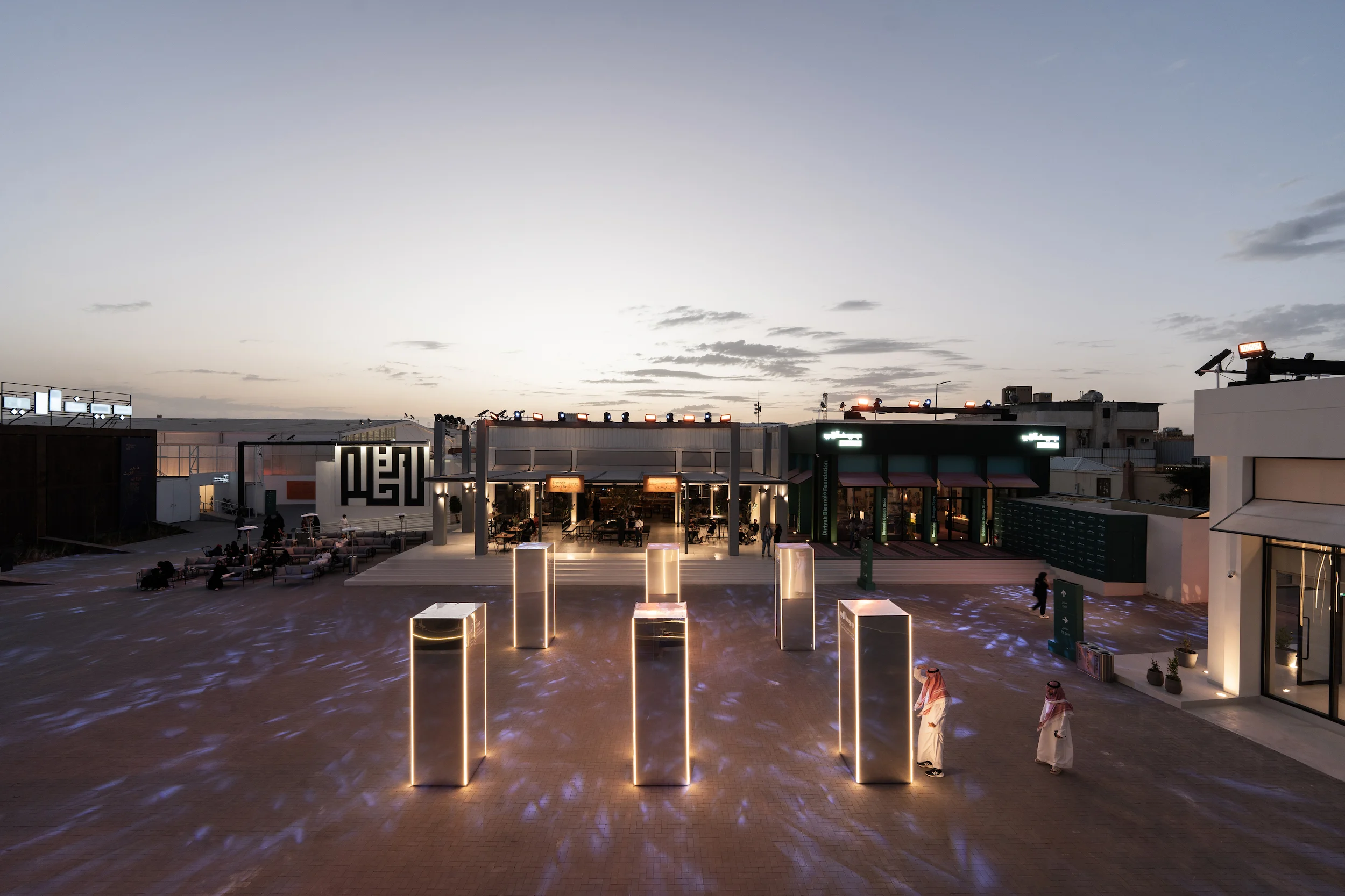 Biennale plaza at dusk with illuminated mirrored totems, new pavilions and a restaurant in the background – Diriyah Contemporary Art Biennale, Riyadh, Saudi Arabia, 2024 – architecture