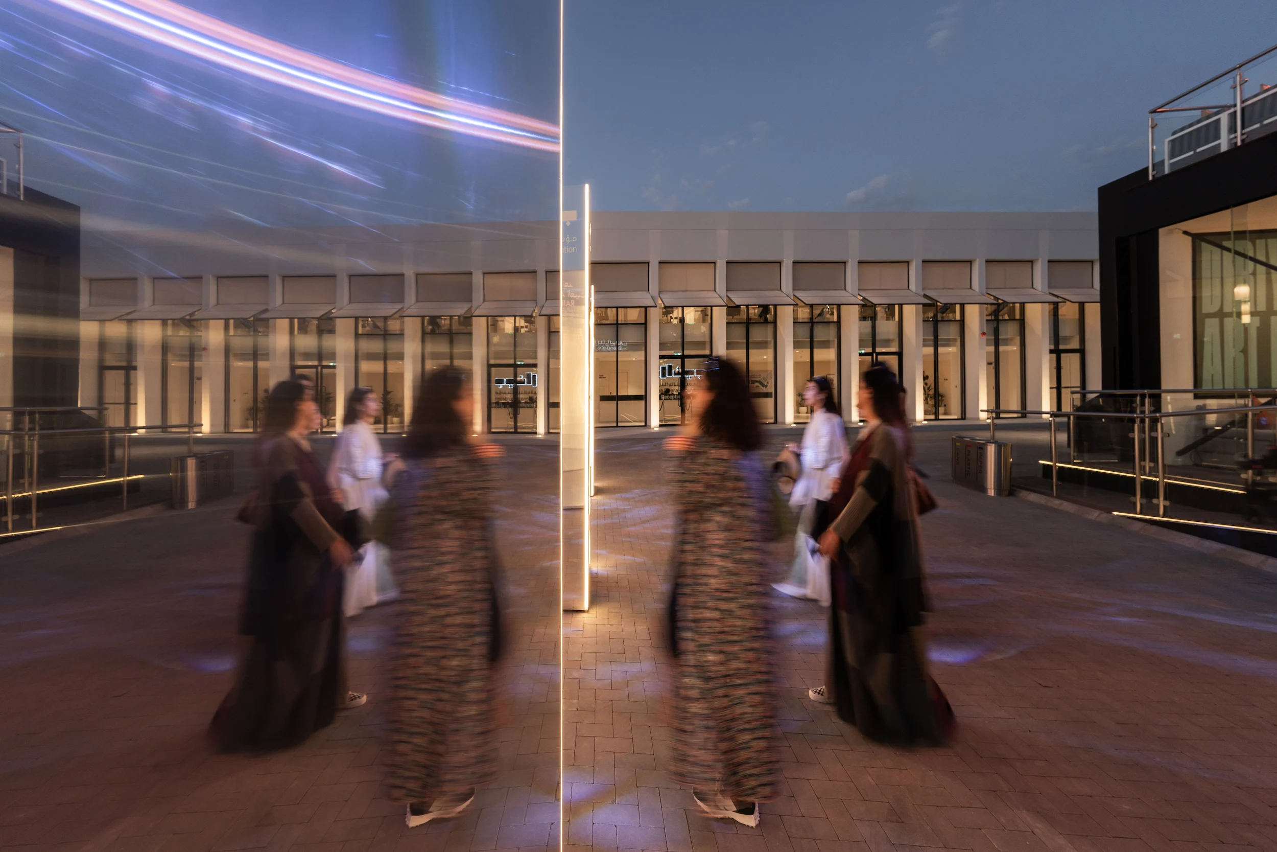 Visitors reflected in a mirrored totem on the plaza at dusk, lit pavilions behind – Diriyah Contemporary Art Biennale, Riyadh, Saudi Arabia, 2024 – architecture