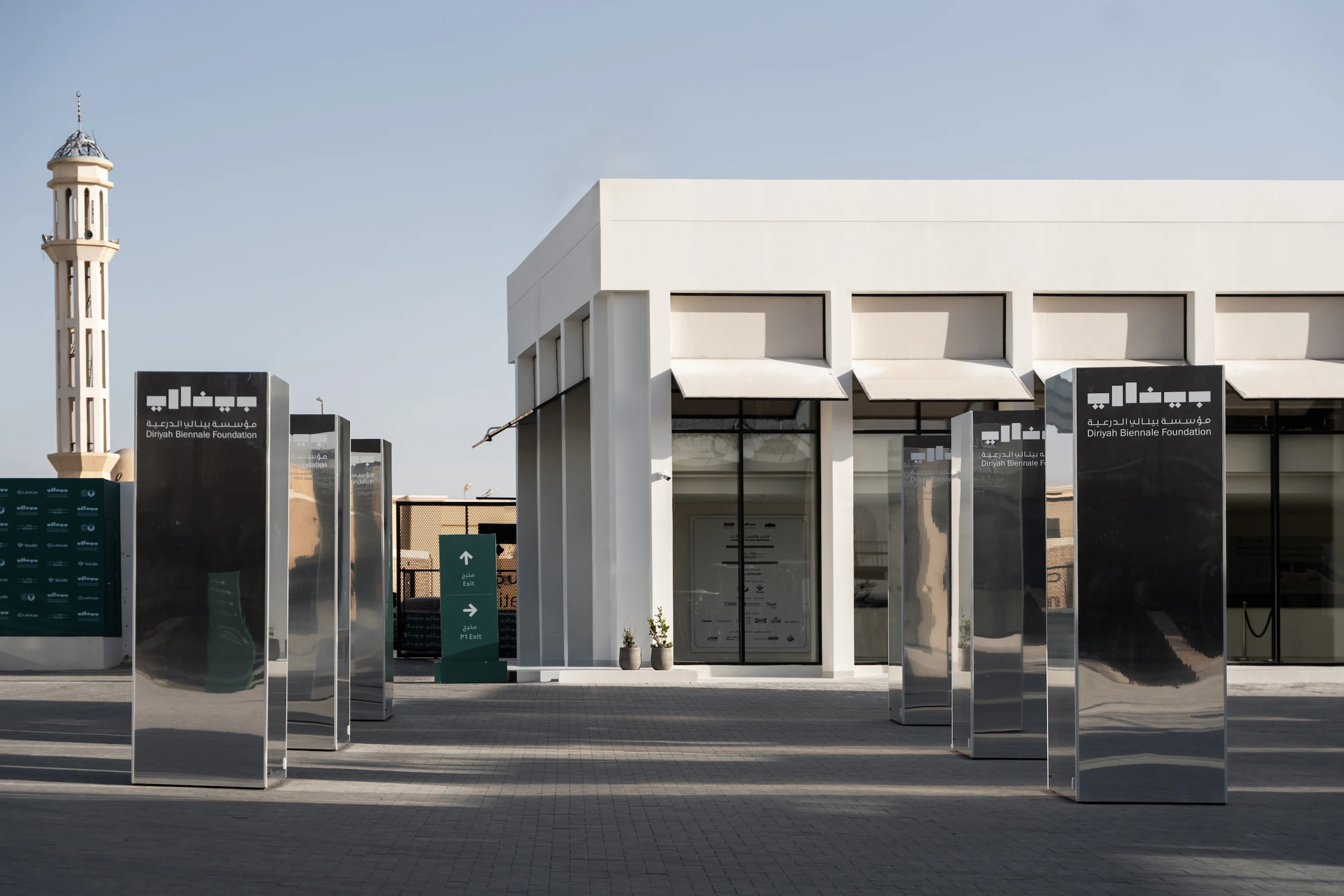 Mirrored steel totems with the Biennale Foundation logo before the white reception building, minaret behind – Diriyah Contemporary Art Biennale, Riyadh, Saudi Arabia, 2024 – architecture