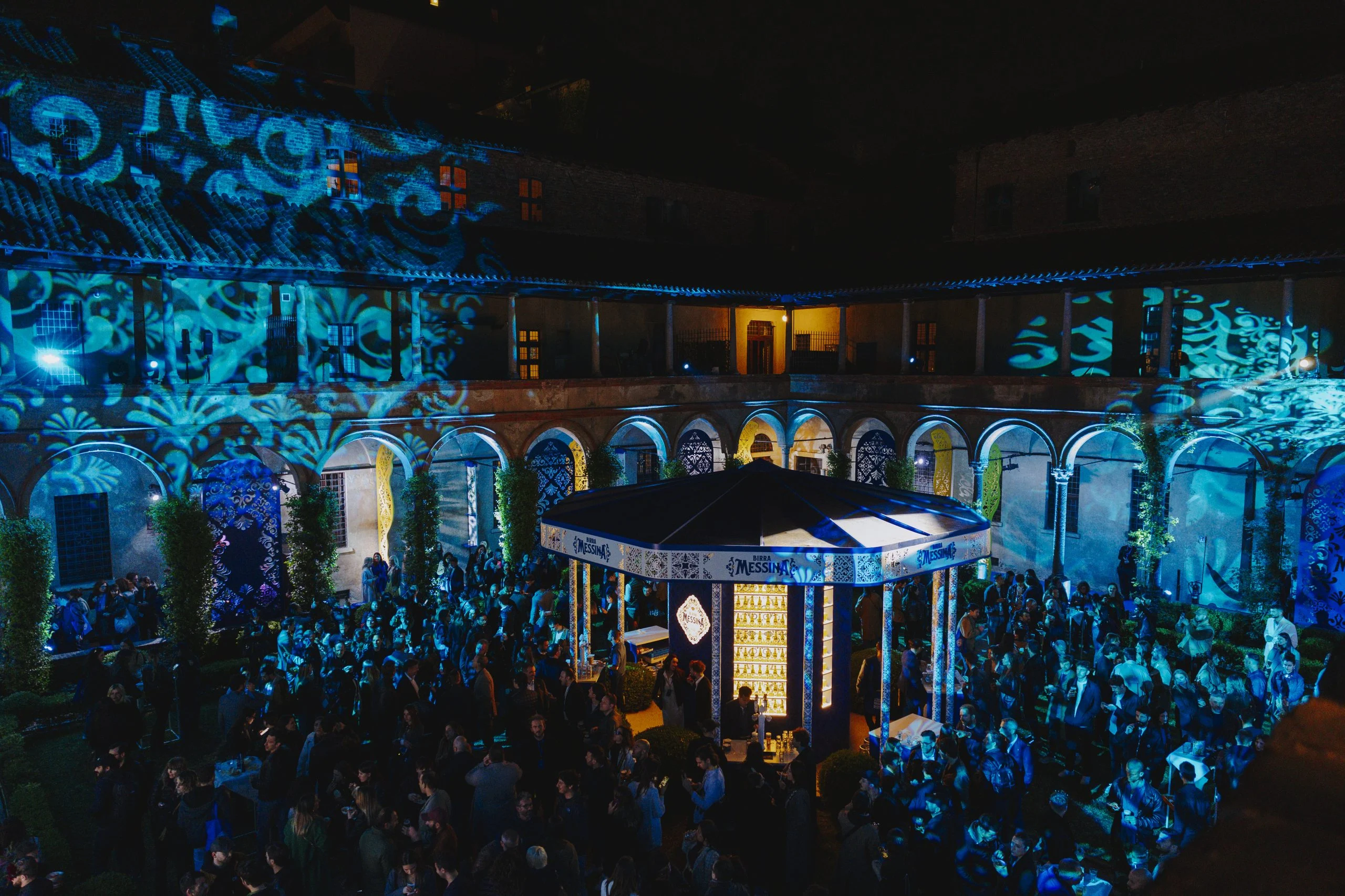 Night view of the crowd around the kiosk, cloister facades covered in blue and turquoise projections – Birra Messina Bar Meraviglia at Fuorisalone, Milan, Italy, 2024 – event design