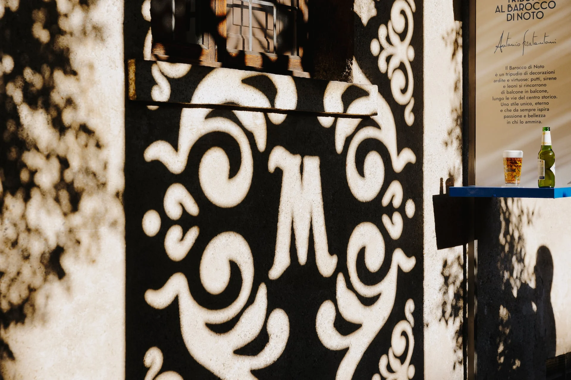 Shadow of a cut-out M monogram cast on a wall beside a shelf holding a beer glass and bottle – Birra Messina Bar Meraviglia at Fuorisalone, Milan, Italy, 2024 – event design