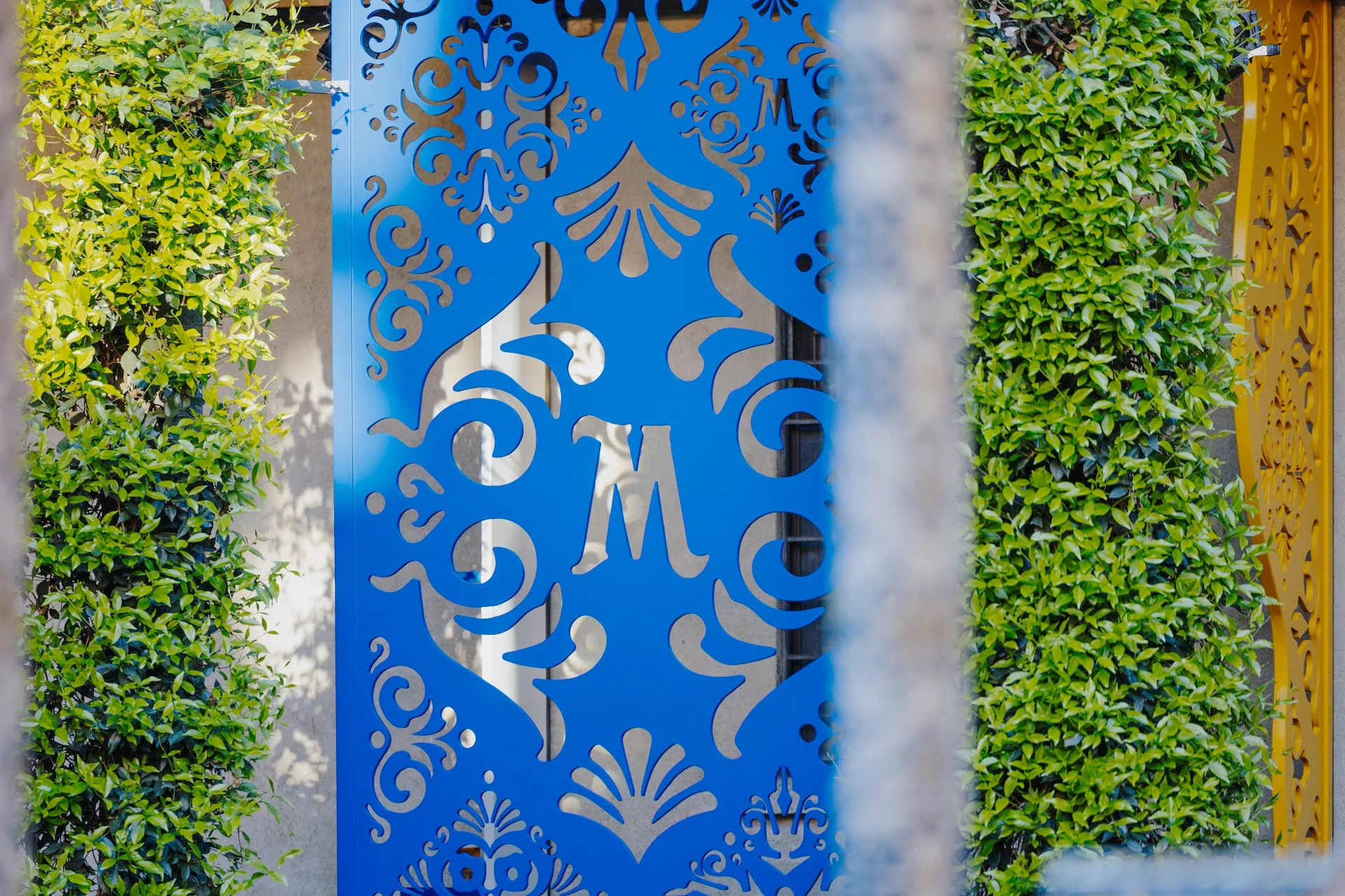 Detail of a blue cut-out metal panel with an M monogram between two green hedges – Birra Messina Bar Meraviglia at Fuorisalone, Milan, Italy, 2024 – event design