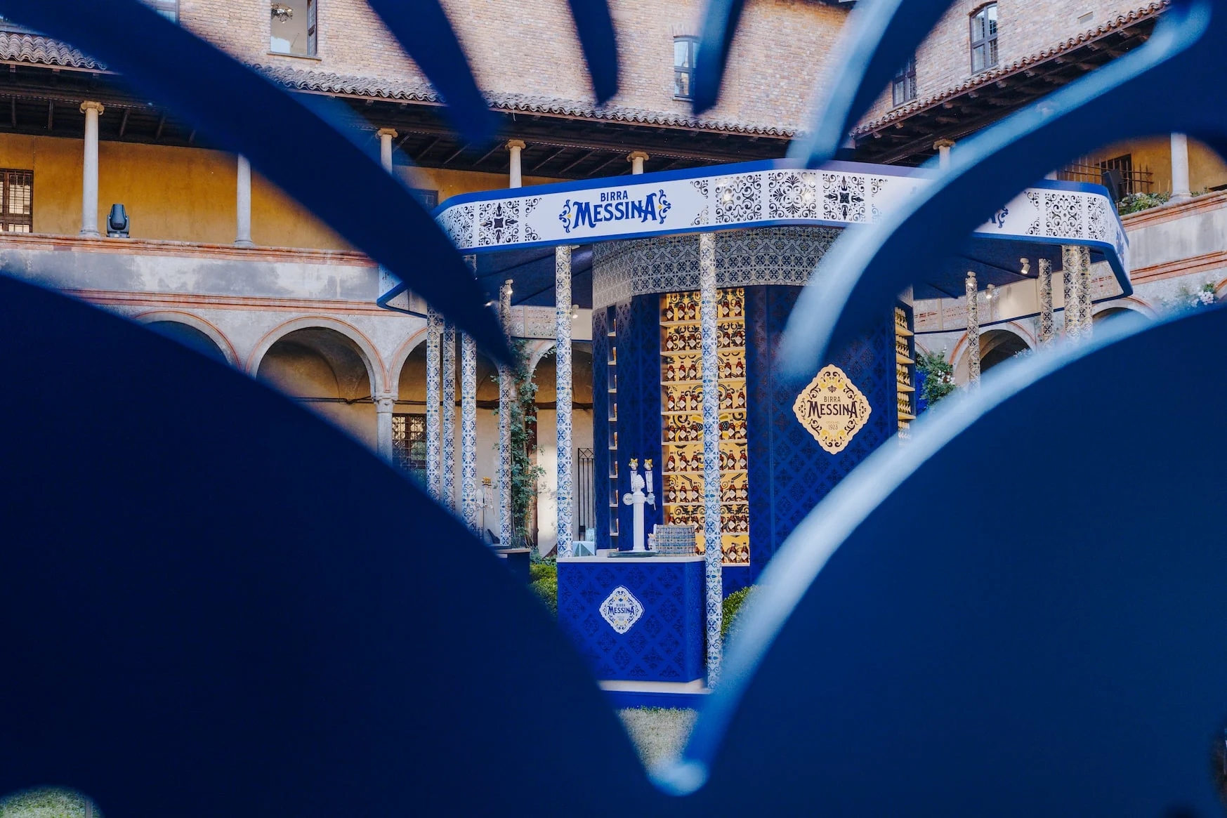 The Birra Messina kiosk framed through blue ornamental cut-out shapes in the foreground – Birra Messina Bar Meraviglia at Fuorisalone, Milan, Italy, 2024 – event design