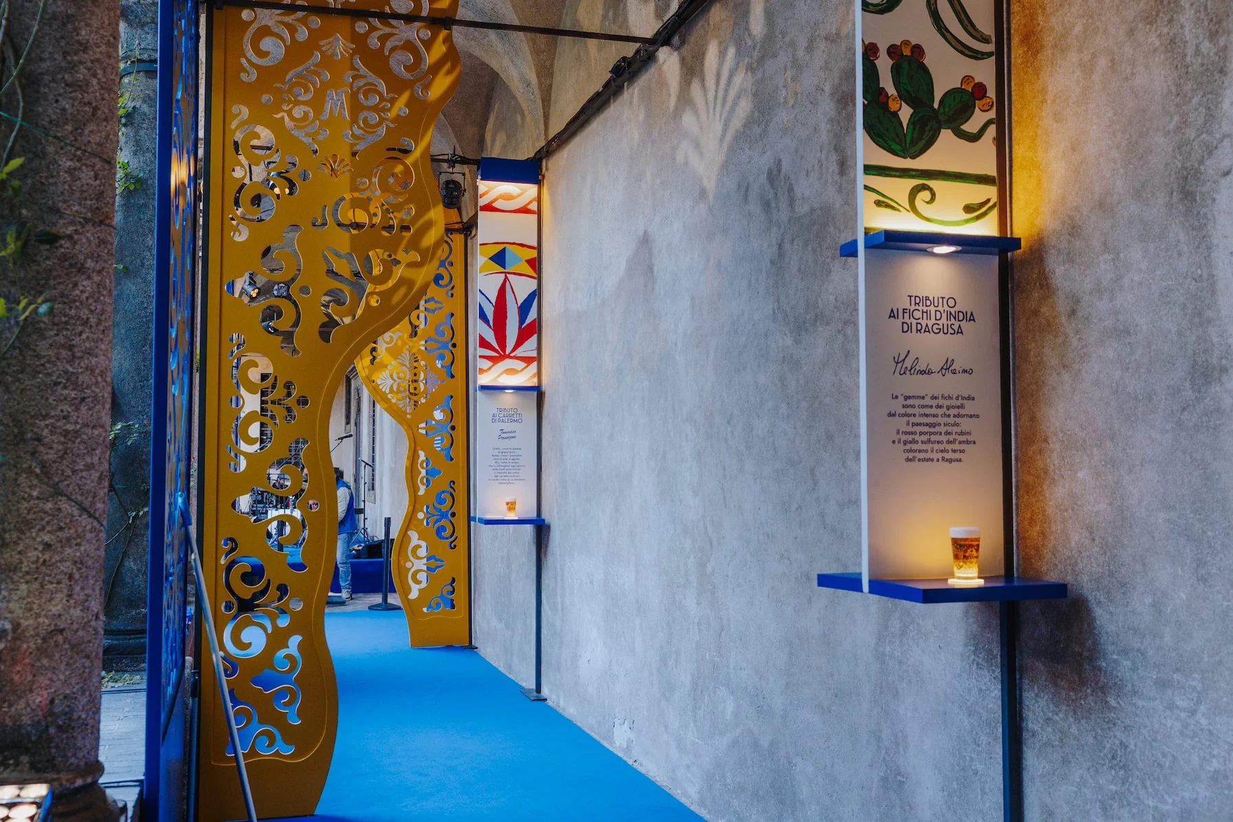 Arcade walkway with golden cut-out panels, blue carpet and illustrated display panels with a glass of beer – Birra Messina Bar Meraviglia at Fuorisalone, Milan, Italy, 2024 – event design