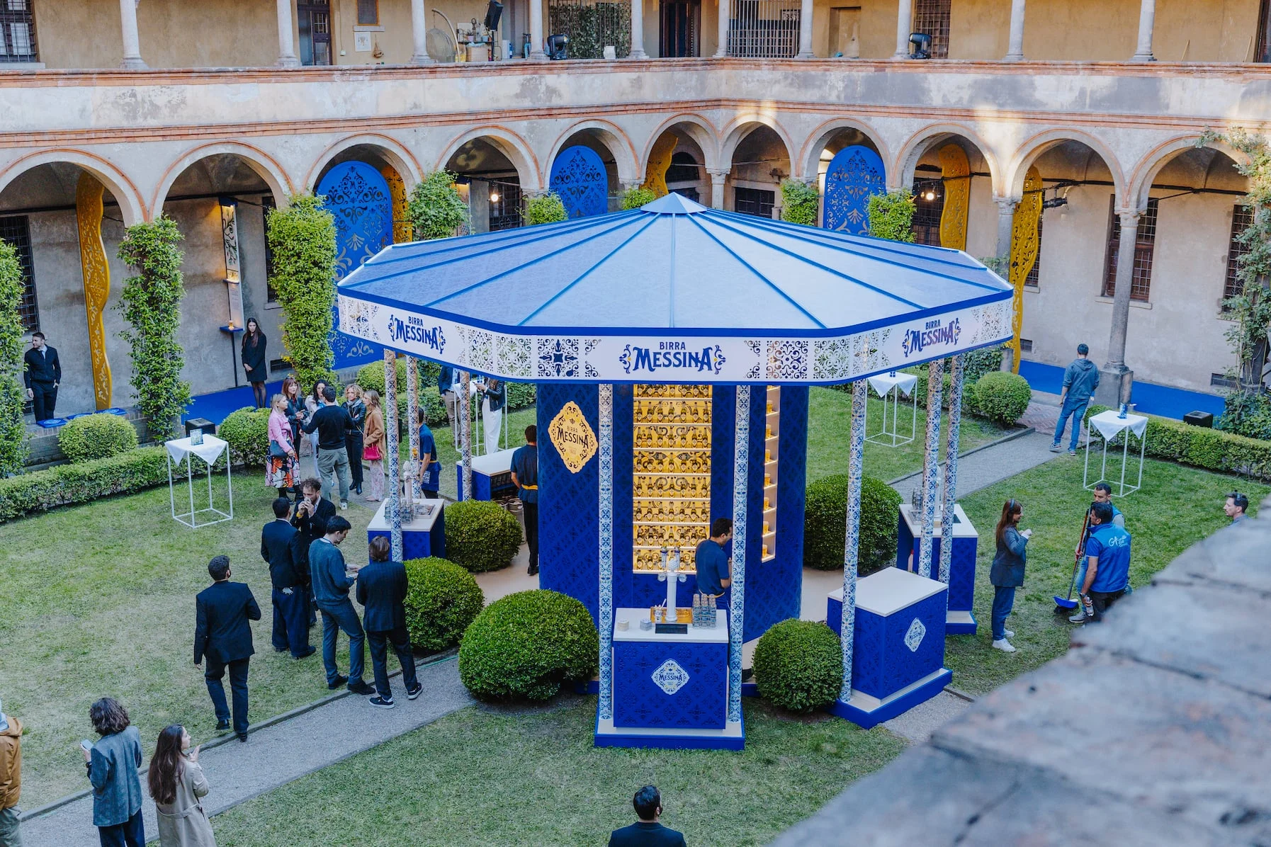 Elevated daytime view of the blue Birra Messina kiosk in the cloister garden, with majolica-style columns – Birra Messina Bar Meraviglia at Fuorisalone, Milan, Italy, 2024 – event design