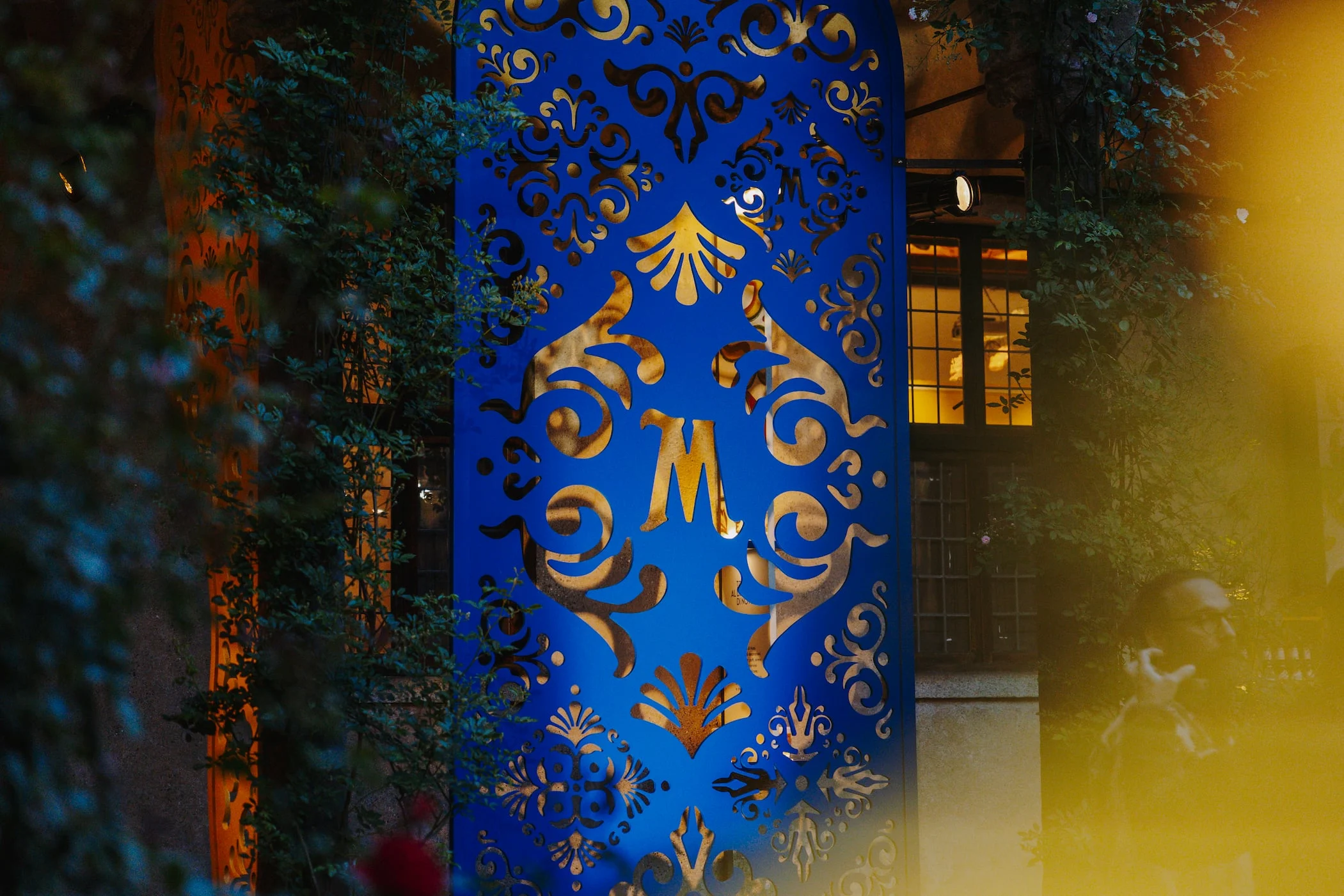 Blue cut-out panel with a golden M monogram glowing at night between dark foliage – Birra Messina Bar Meraviglia at Fuorisalone, Milan, Italy, 2024 – event design